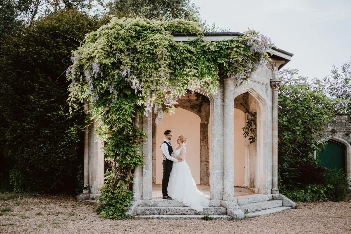 Wedding couple in wedding dress and tuxedo standing in a stone pavilion surrounded by lush greenery and flowering vines.