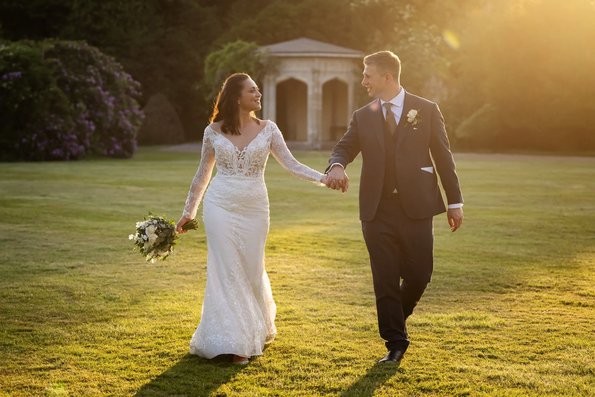 A bride and groom walking hand in hand on a grassy field during sunset, with trees and a small structure in the background.
