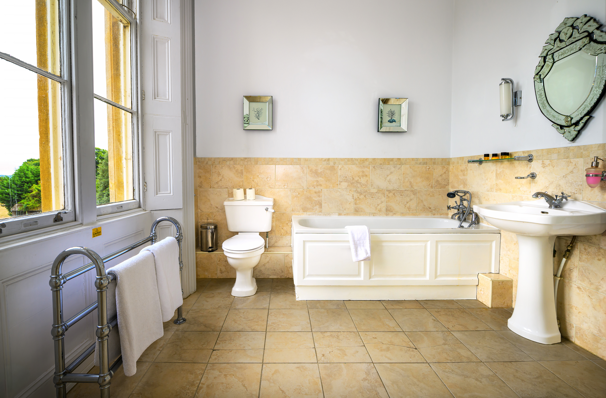 A bathroom with beige tiled floors and half tiled walls, a large window with open blinds, a white toilet, a white bathtub with a towel hanging over its side, a white pedestal sink, a decorative mirror, framed pictures of plants, wall-mounted shelves,