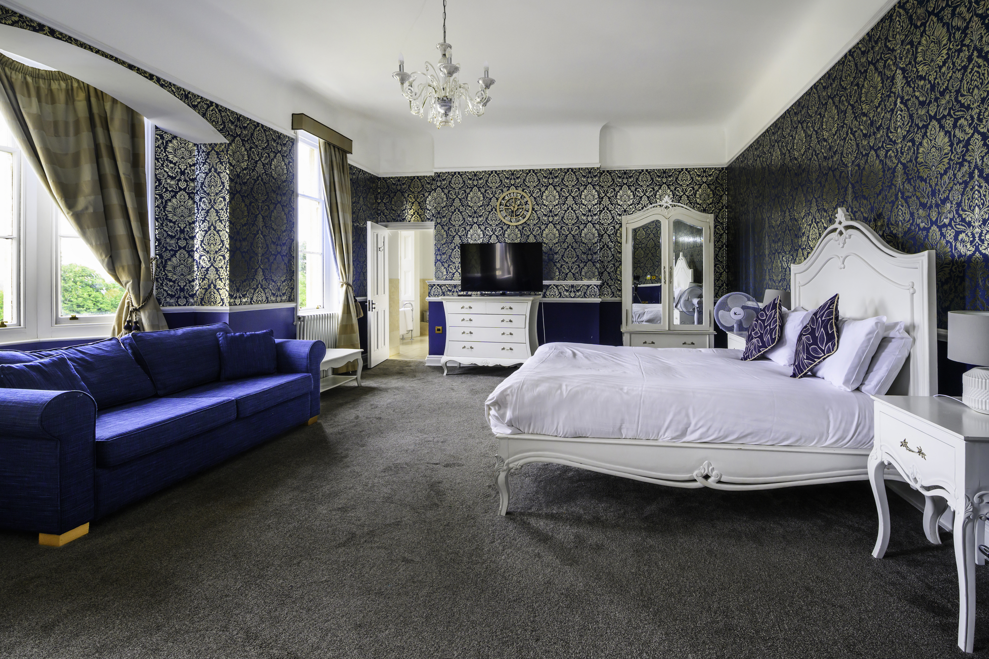 A spacious bedroom featuring a large white bed with decorative pillows, a blue velvet sofa, a white dresser with a TV, a white wardrobe with a mirror, and three windows with gold curtains, all set against dark, patterned wallpaper.