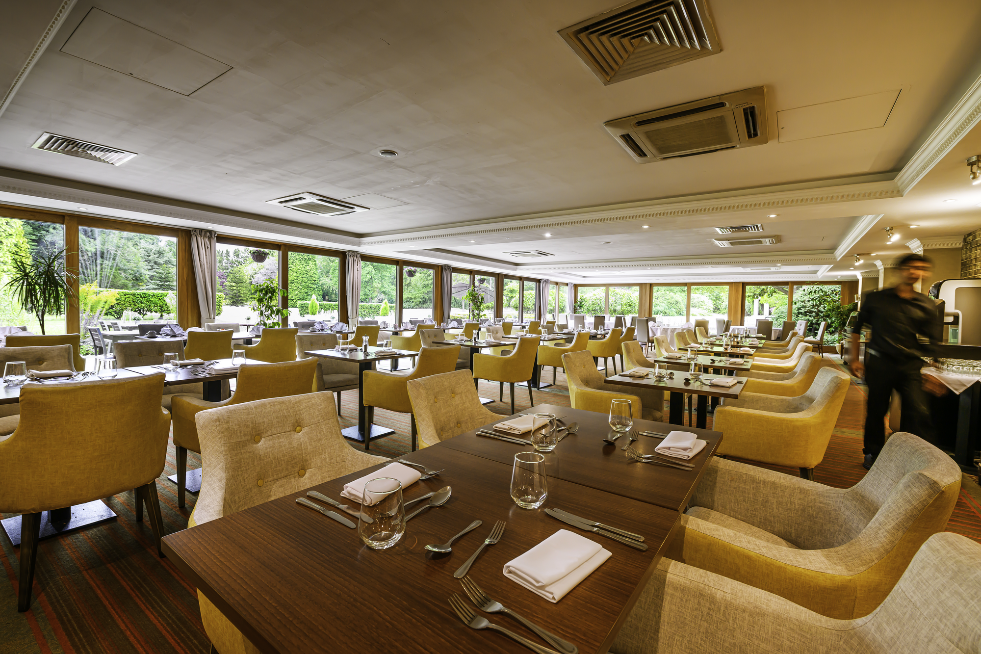 Elegant restaurant dining area with tables set with glassware, silverware, and napkins, surrounded by cream and yellow upholstered chairs, large windows overlooking a lush garden, and a blurred waiter in the background.