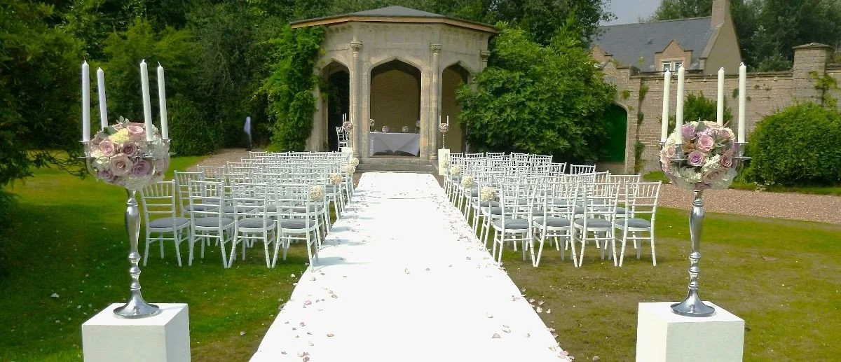 Outdoor wedding ceremony setup with white chairs on grass, an aisle with flower petals, and floral candle holders at the entrance, in front of a gazebo surrounded by trees and greenery.
