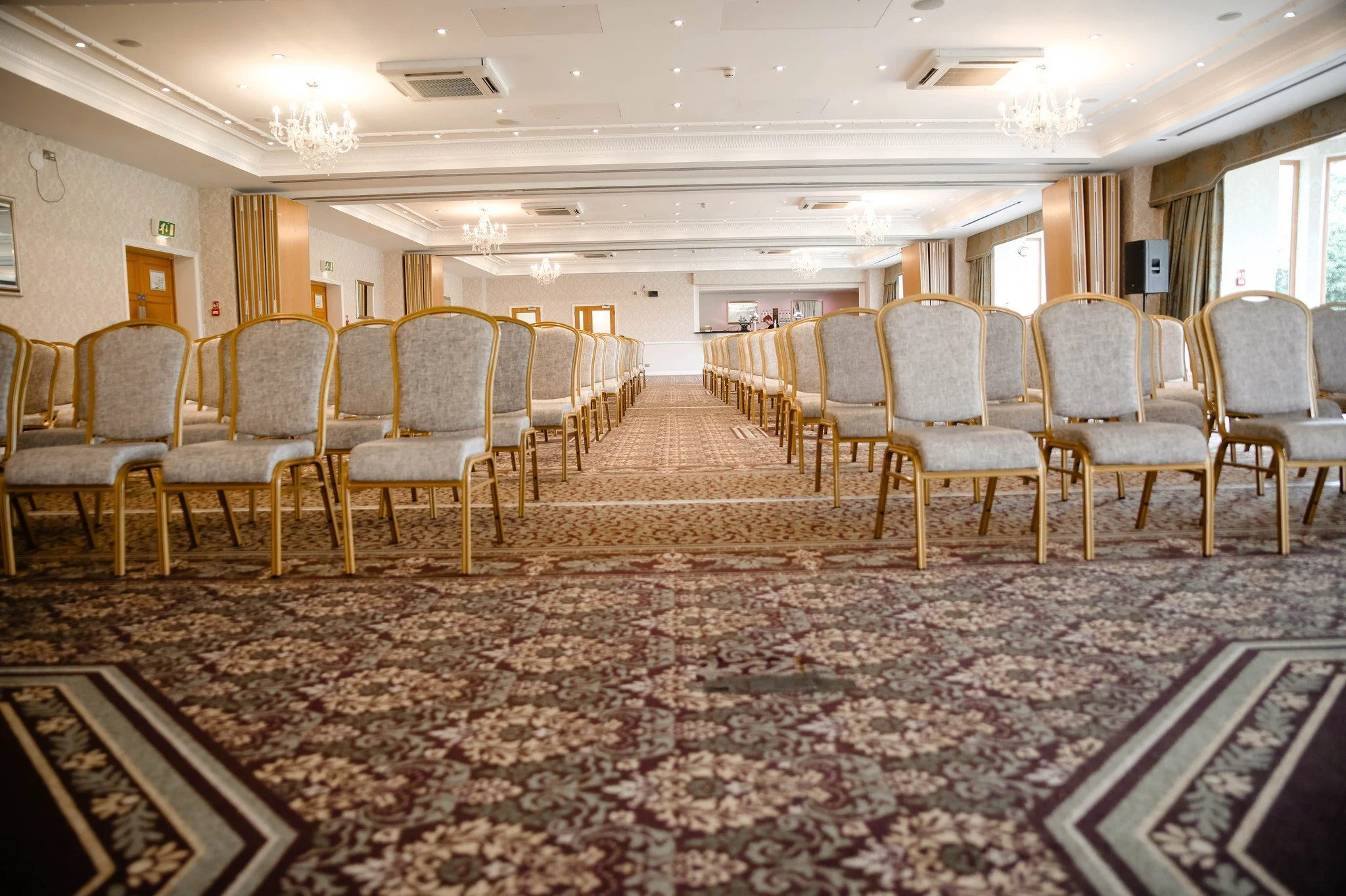Conference room with rows of beige chairs, chandeliers, and large windows letting in natural light.