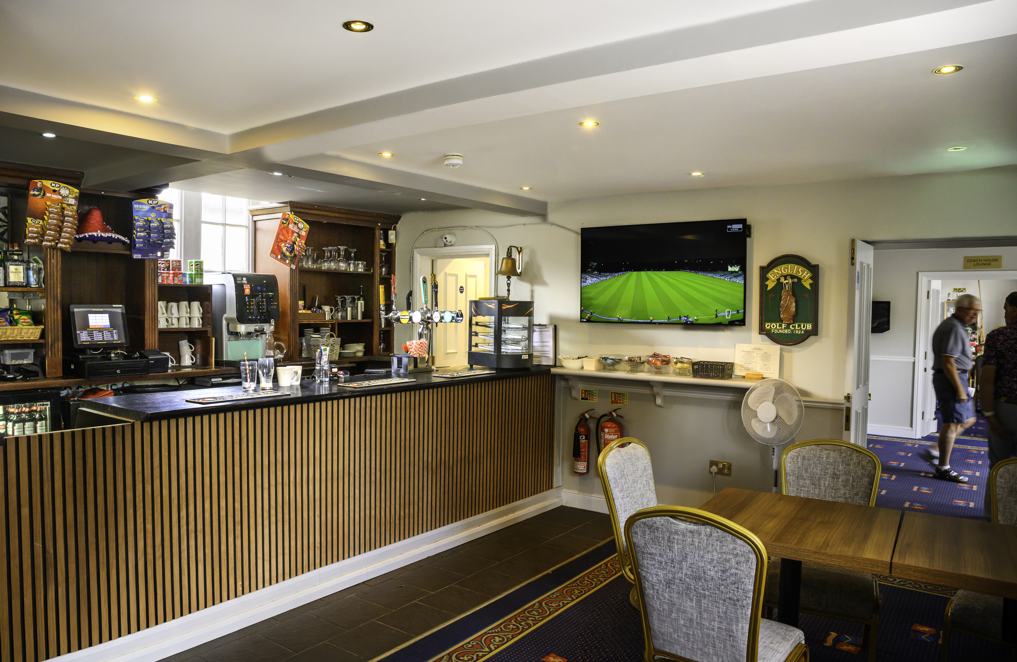 Indoor lounge area of a golf club with a bar, TV playing a golf game, and people walking in the background.