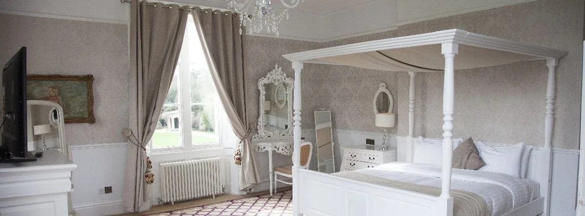 Bedroom with a white four-poster bed, a window with cream curtains, a dresser with a mirror, a TV on a white cabinet, and a small white side table with a lamp.