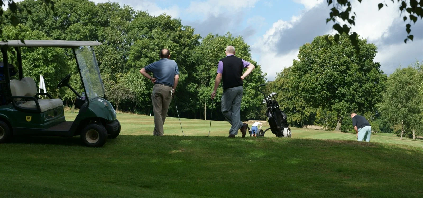Two men standing on a golf course with their golf clubs, observing their game, while another man is putting on the grass in the background. A golf cart is parked to the left.