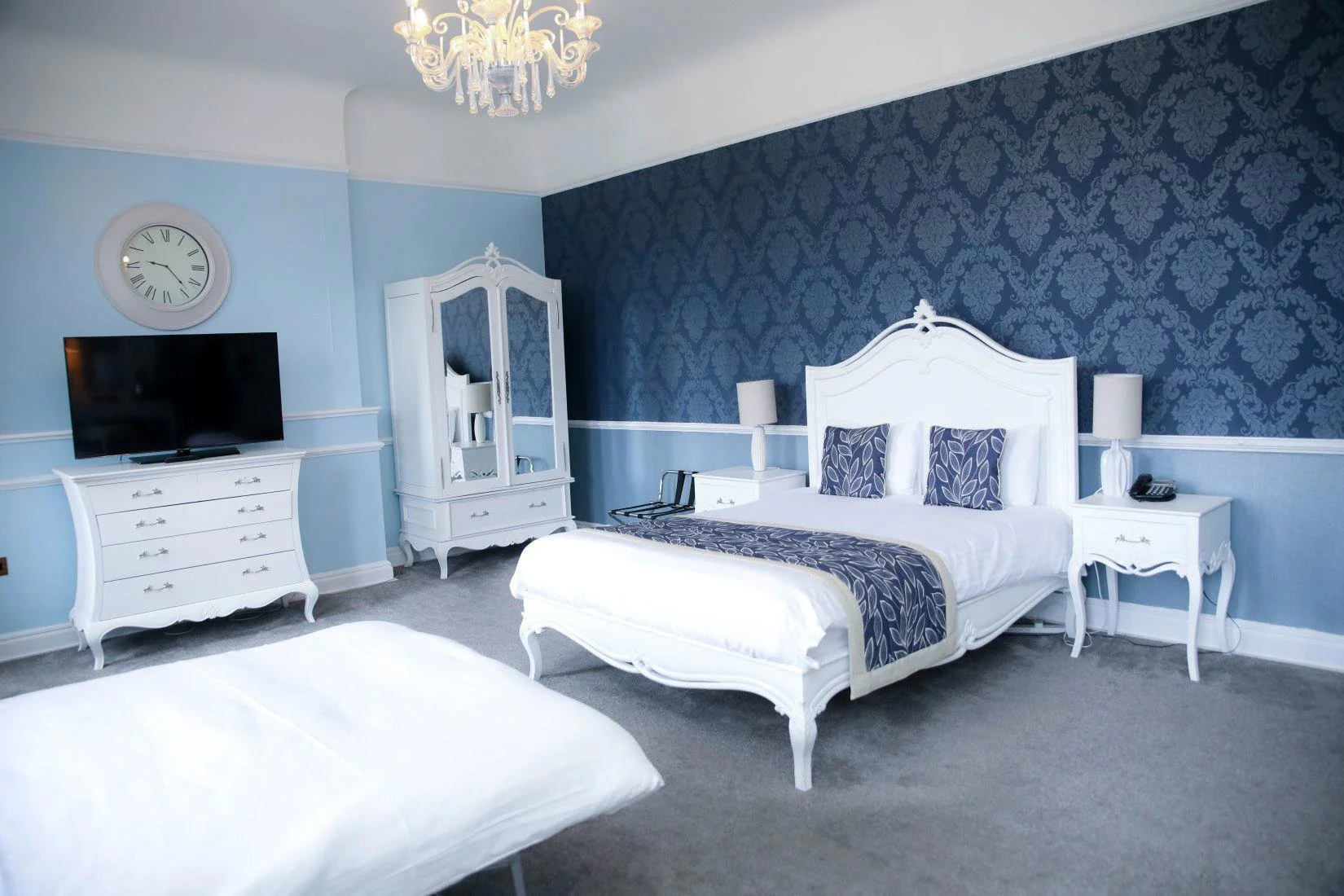 A bedroom with light blue walls and darker blue wallpapered accent wall behind the bed. The room features a white bed with matching nightstands, a white dresser with a TV on top, a white wardrobe with mirrored doors, and a chandelier hanging from the