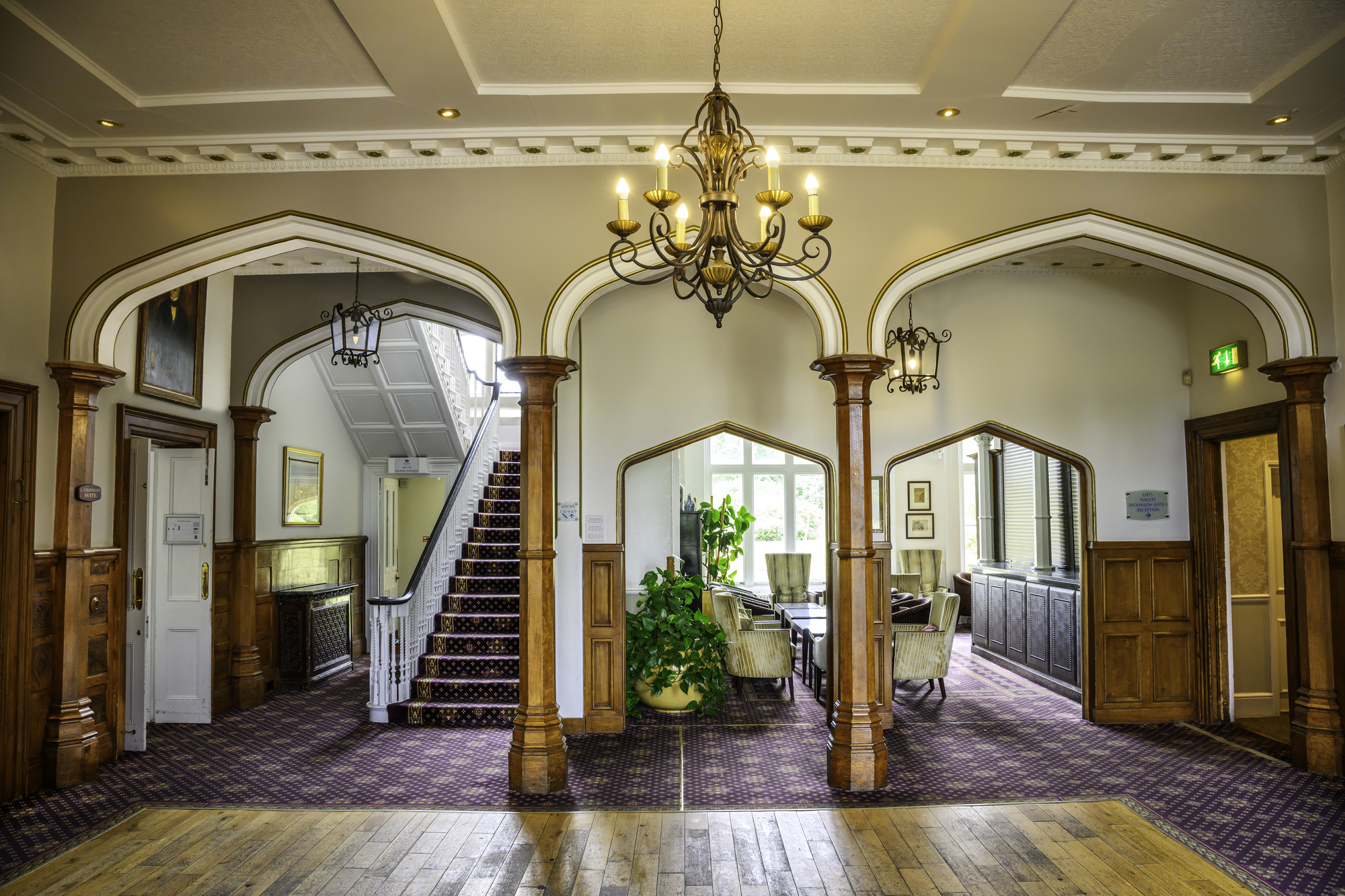 Interior of a vintage-style building with wooden archways, a chandelier, and a staircase. There are sitting areas with chairs and plants near windows.