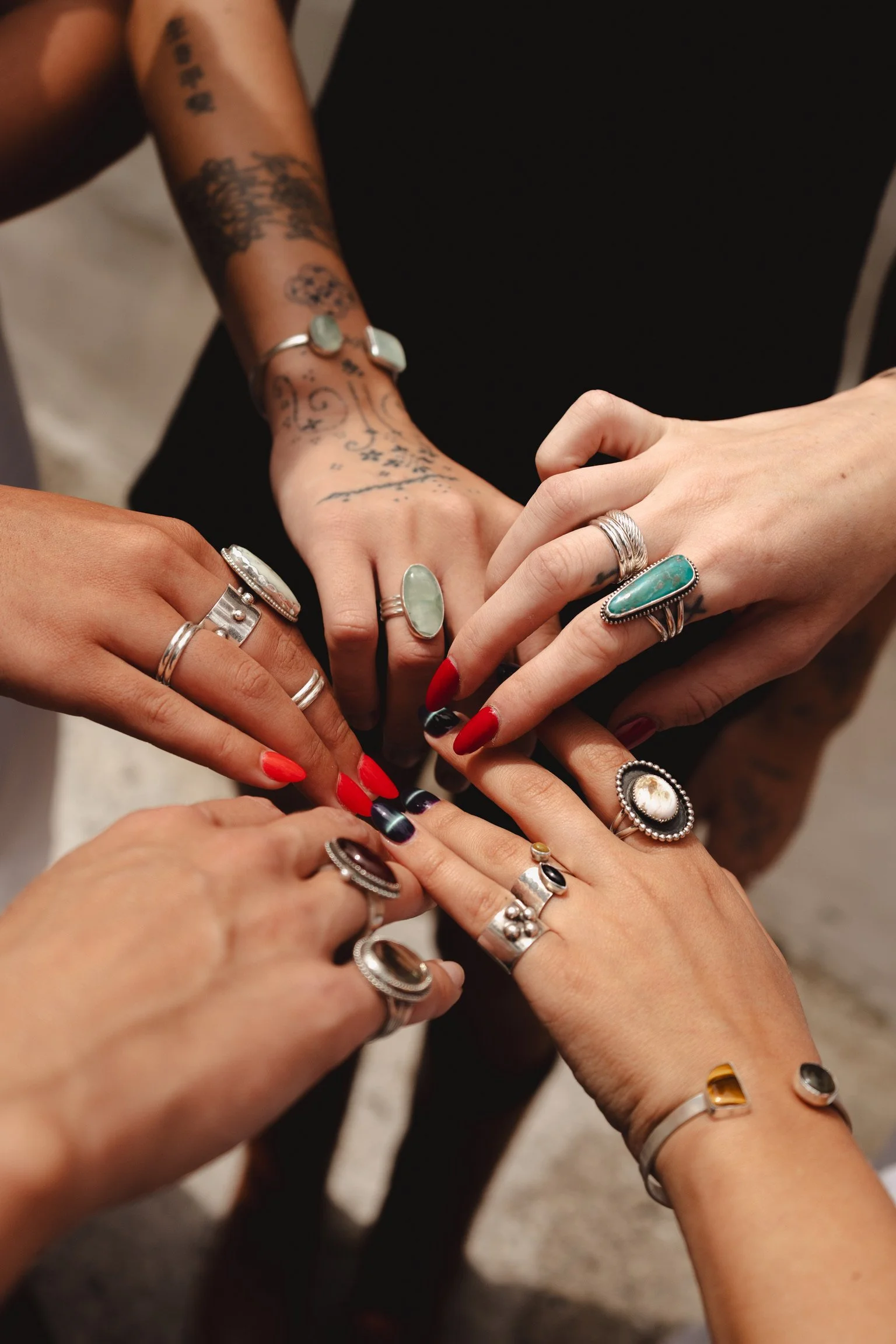 Multiple hands adorned with rings, displaying tattoos, and having painted nails, are overlapping in a gesture of unity.