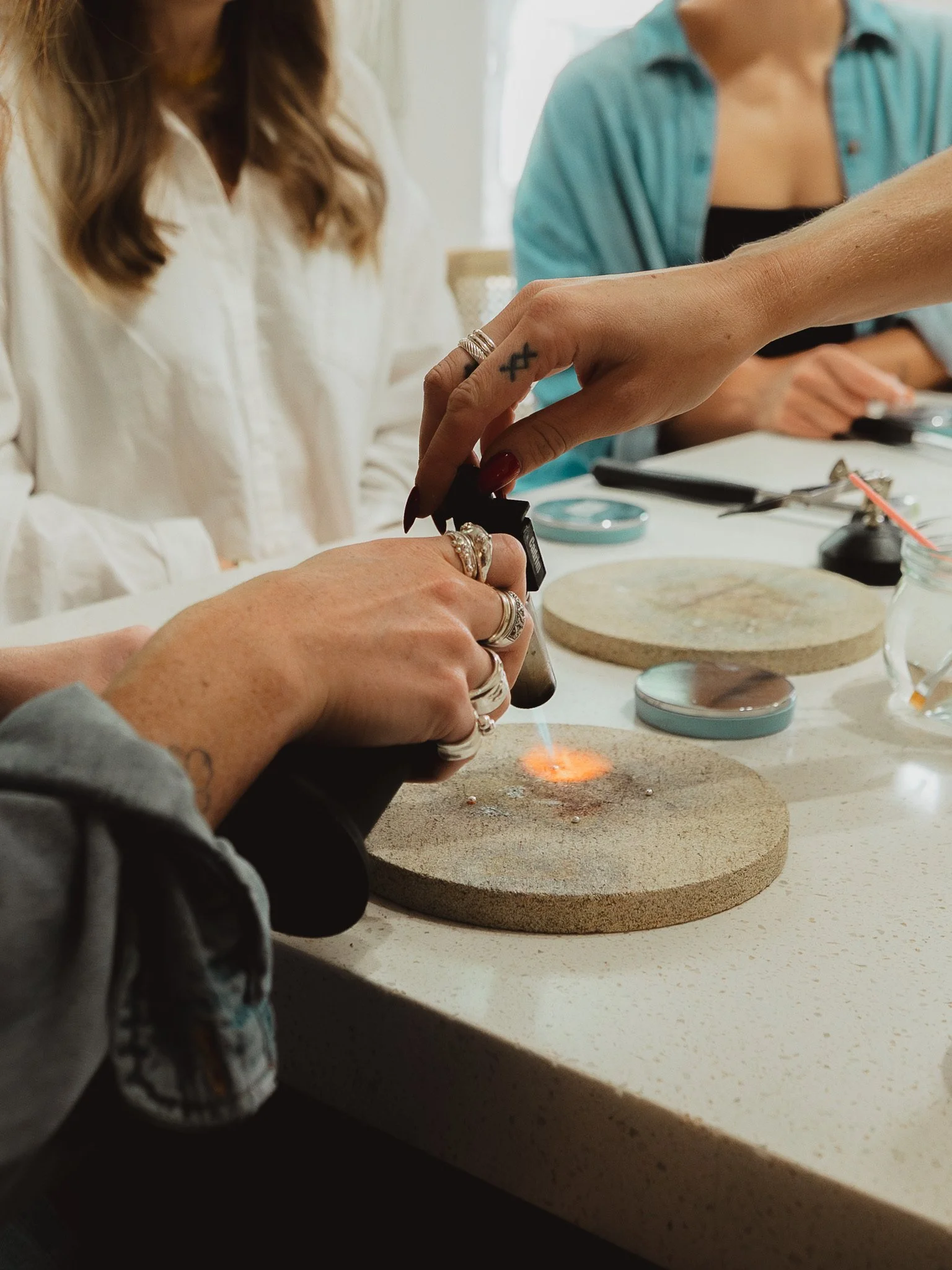 A person participating in a silversmithing workshop, holding a torch and soldering her jewelry piece together with the assistance of a silversmith instructor