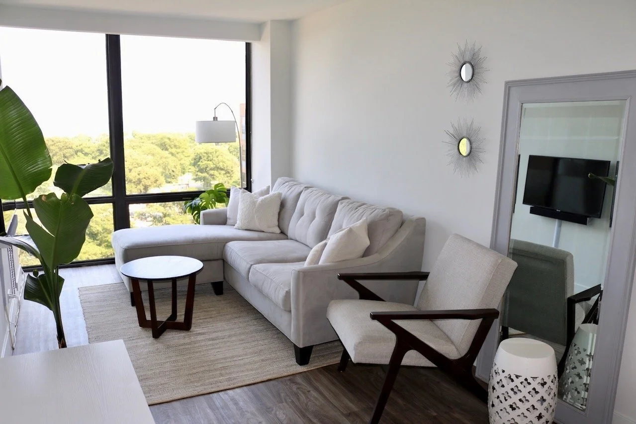 Modern living room with large window, white sectional sofa, beige armchair, mirrors, TV, and indoor plants.