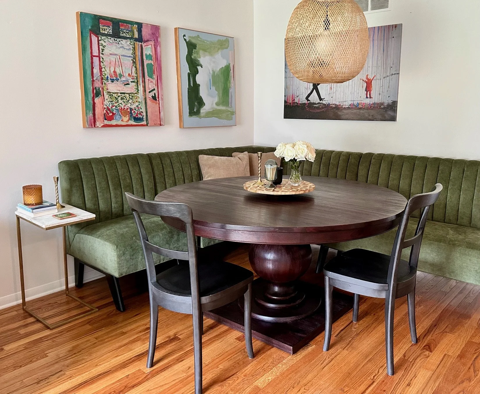 A dining area with a round wooden table, surrounded by a green upholstered bench and three black chairs. The table has a vase with white flowers. On the left side, a small white side table with books and a candle. The wall behind has three colorful a