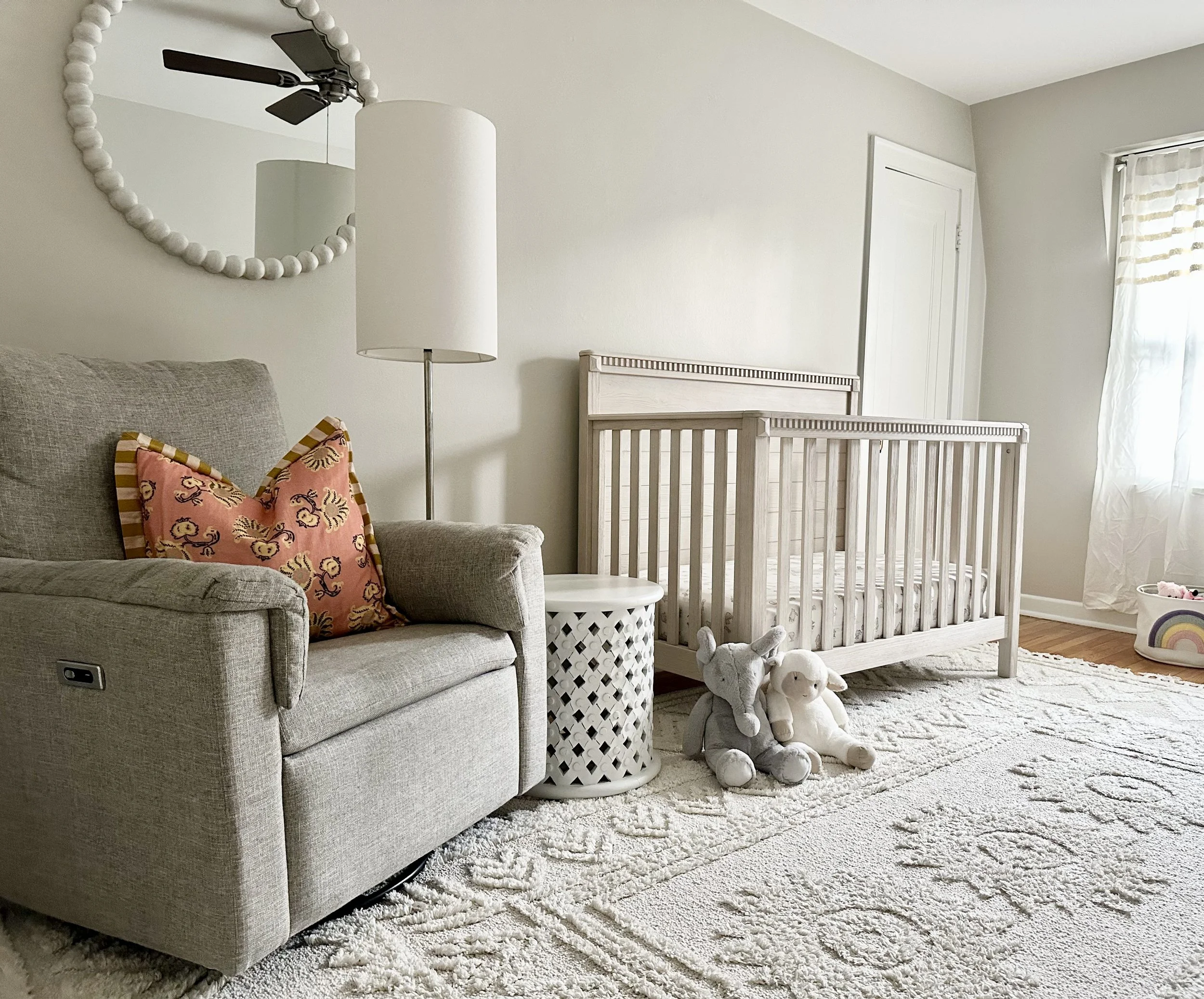 A cozy nursery room with a beige sofa and an orange and patterned pillow, a white floor lamp, a wooden crib, stuffed animals, a white patterned side table, a round mirror, a window with sheer curtains, and a cream-colored patterned rug.