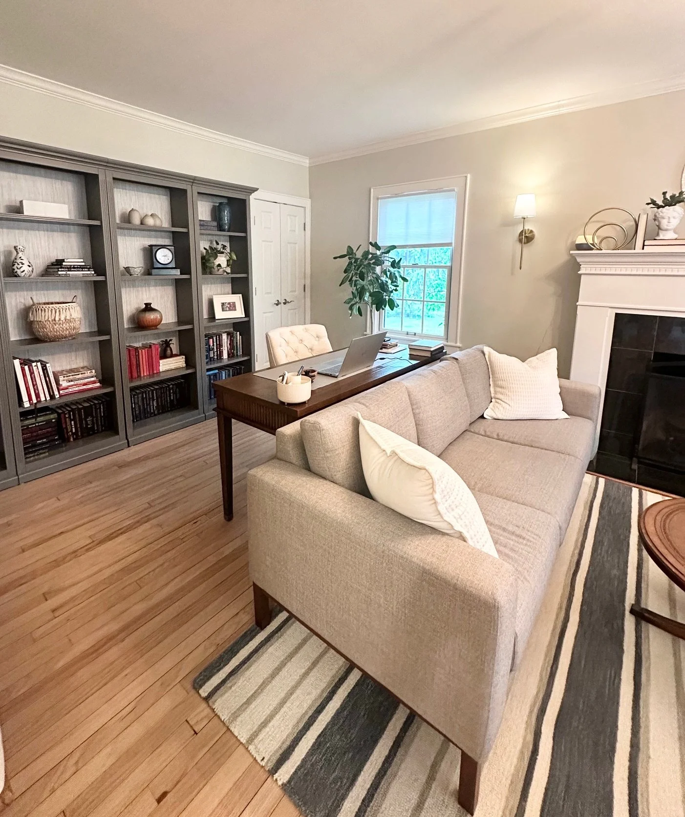 Living room with beige sofa, wooden desk, bookshelves, and a window with greenery outside.