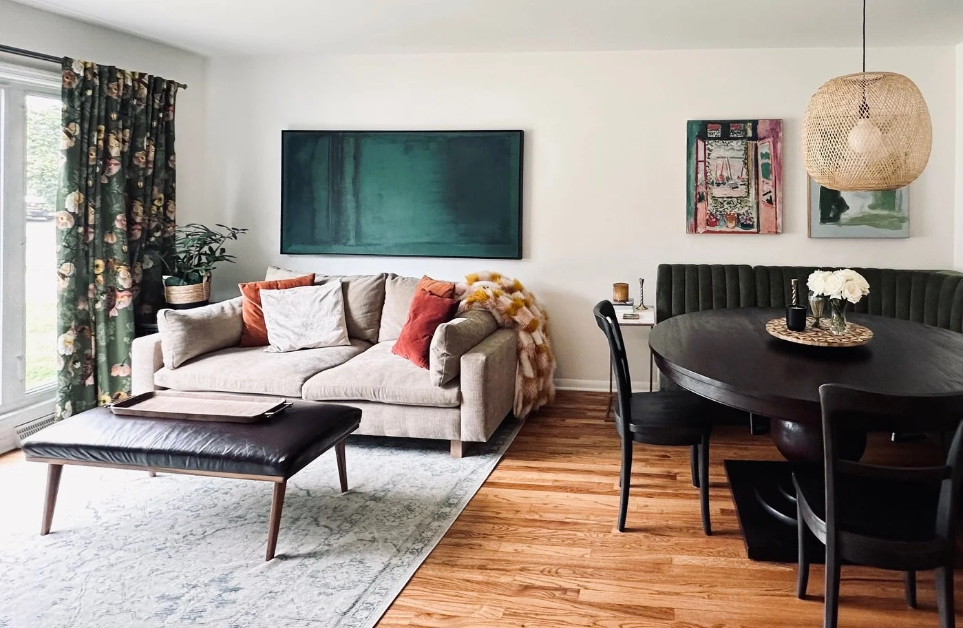 A living room with a beige sofa decorated with throw pillows, a black leather ottoman tray, and a coffee table with a white rug underneath. A large window with floral curtains, a potted plant, and wall art including a large abstract piece and smaller