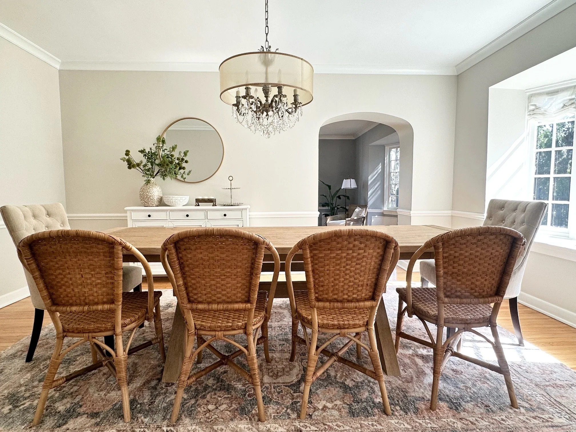 A dining room with six wicker chairs around a wooden table, a white sideboard with a vase, a bowl, and decorative items, a chandelier, and a large window with sunlight.