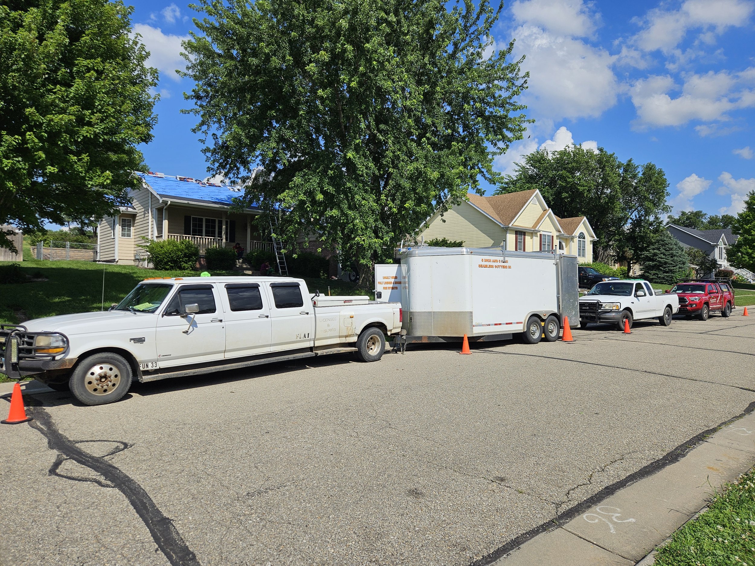 Residential street with a large white utility truck towing a trailer, parked in front of a house under renovation. The house has a blue tarp on the roof. Other vehicles, including a white pickup and red SUV, are parked nearby. Orange traffic cones ar