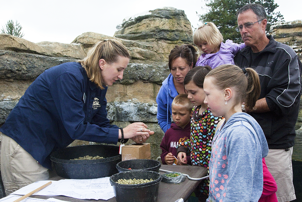 A staff member in a blue shirt demonstrates modeling to a ground of children and adults.
