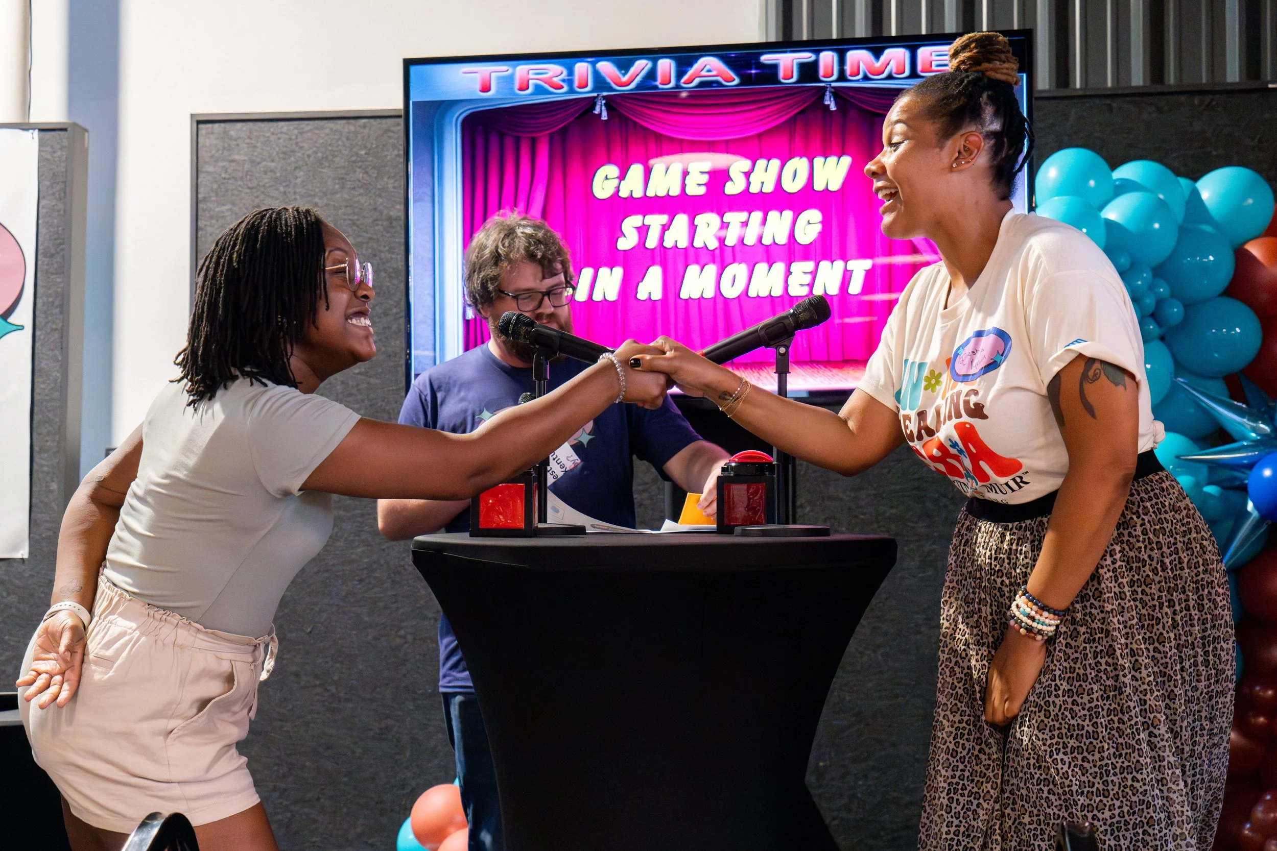 Two women shake hands during a game show at a trivia event, with a large screen in the background displaying 'TRIVIA TIME' and 'GAME SHOW STARTING IN A MOMENT,' in front of balloons and a curtained stage.