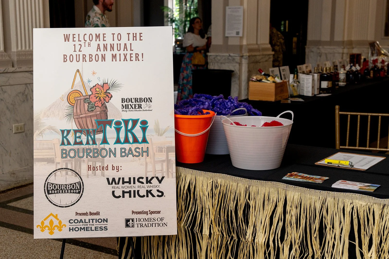 Sign for the 12th annual Kentiki Bourbon Bash event with sponsor logos, next to tables with purple and red candy dishes in an elegant room.