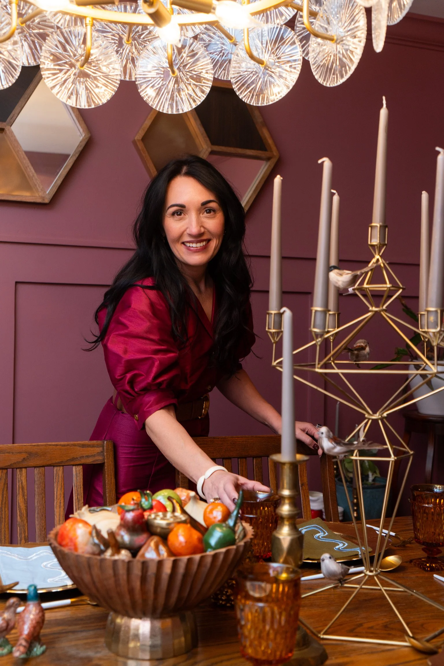 A woman with long dark hair smiling while setting a dining table, surrounded by candles, a bowl of decorative fruit, and bird figurines, in a room with purple walls and hexagonal mirrors.