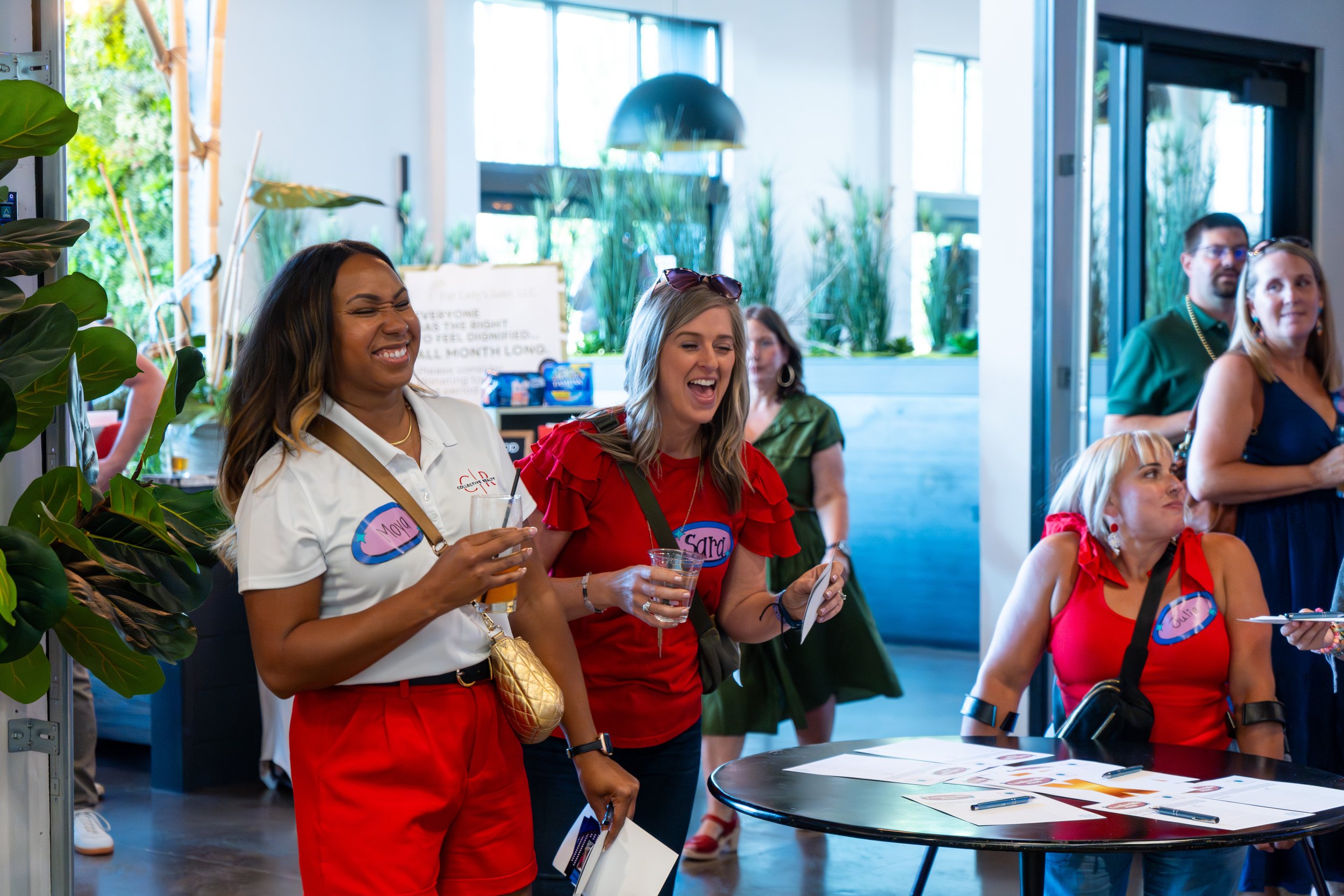 Smiling women at a social event, holding drinks, with name tags, in a brightly lit indoor space with large windows and plants.