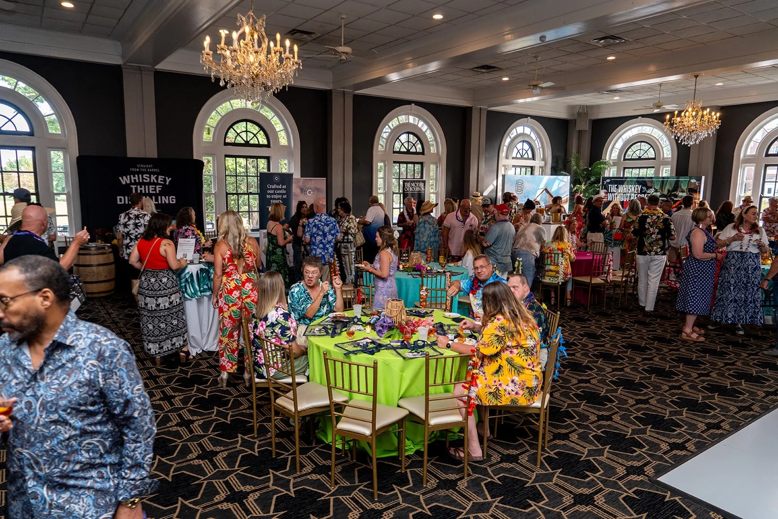 A large non-profit event with people wearing colorful tropical shirts and dresses. There are decorated tables, a chandelier, large windows, and banners promoting whiskey.