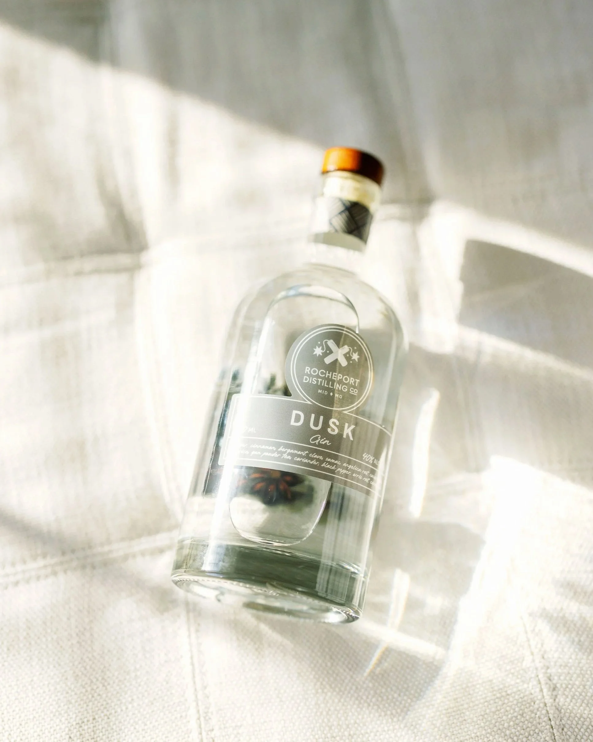 A clear glass bottle of Rochemport Distilling Co.'s Dusk Gin with a cork stopper on a white textured surface, with sunlight creating shadows.