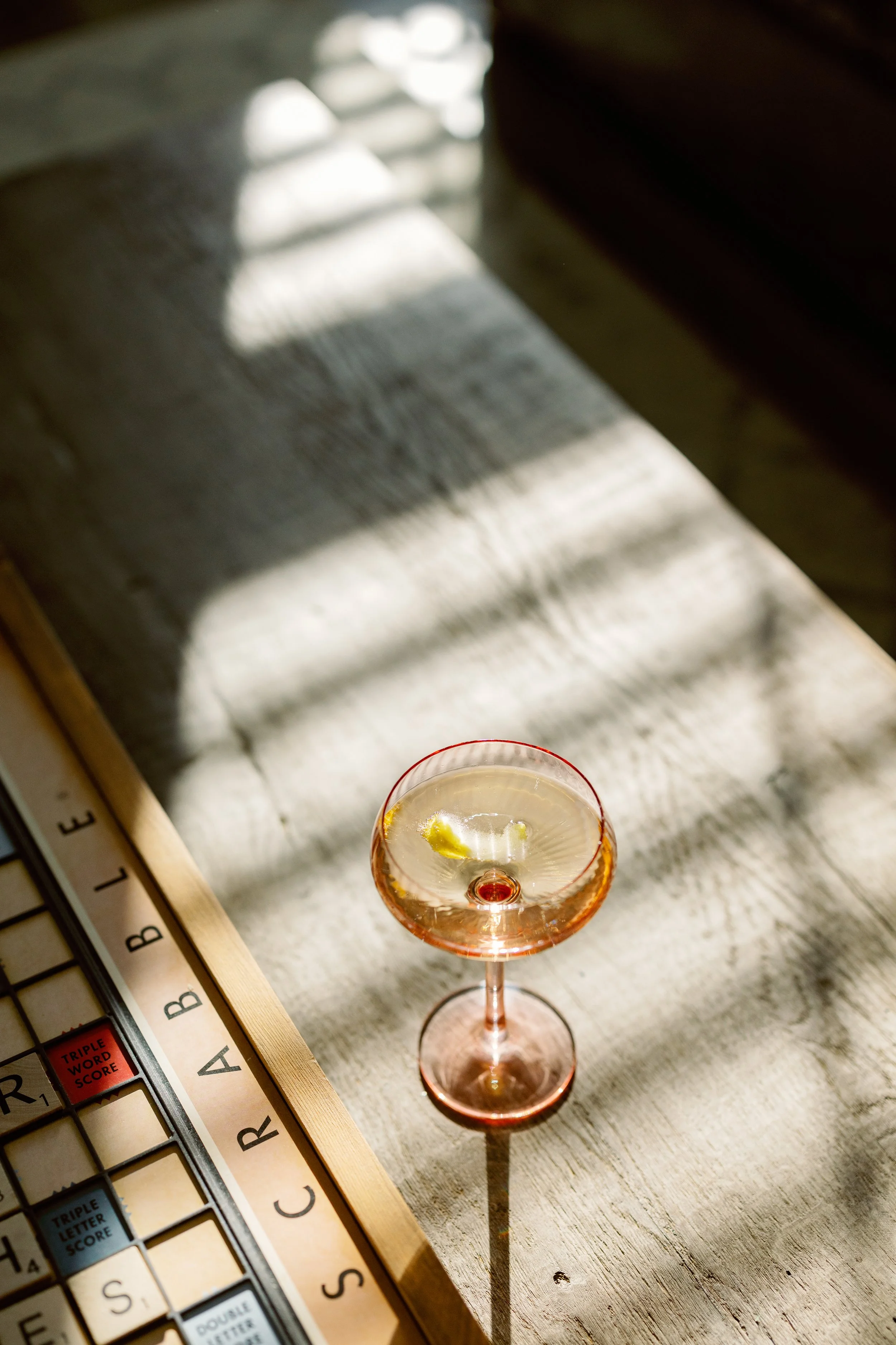 A glass of champagne with a lemon twist, casting a shadow on a rustic wooden table with sunlight and shadows.