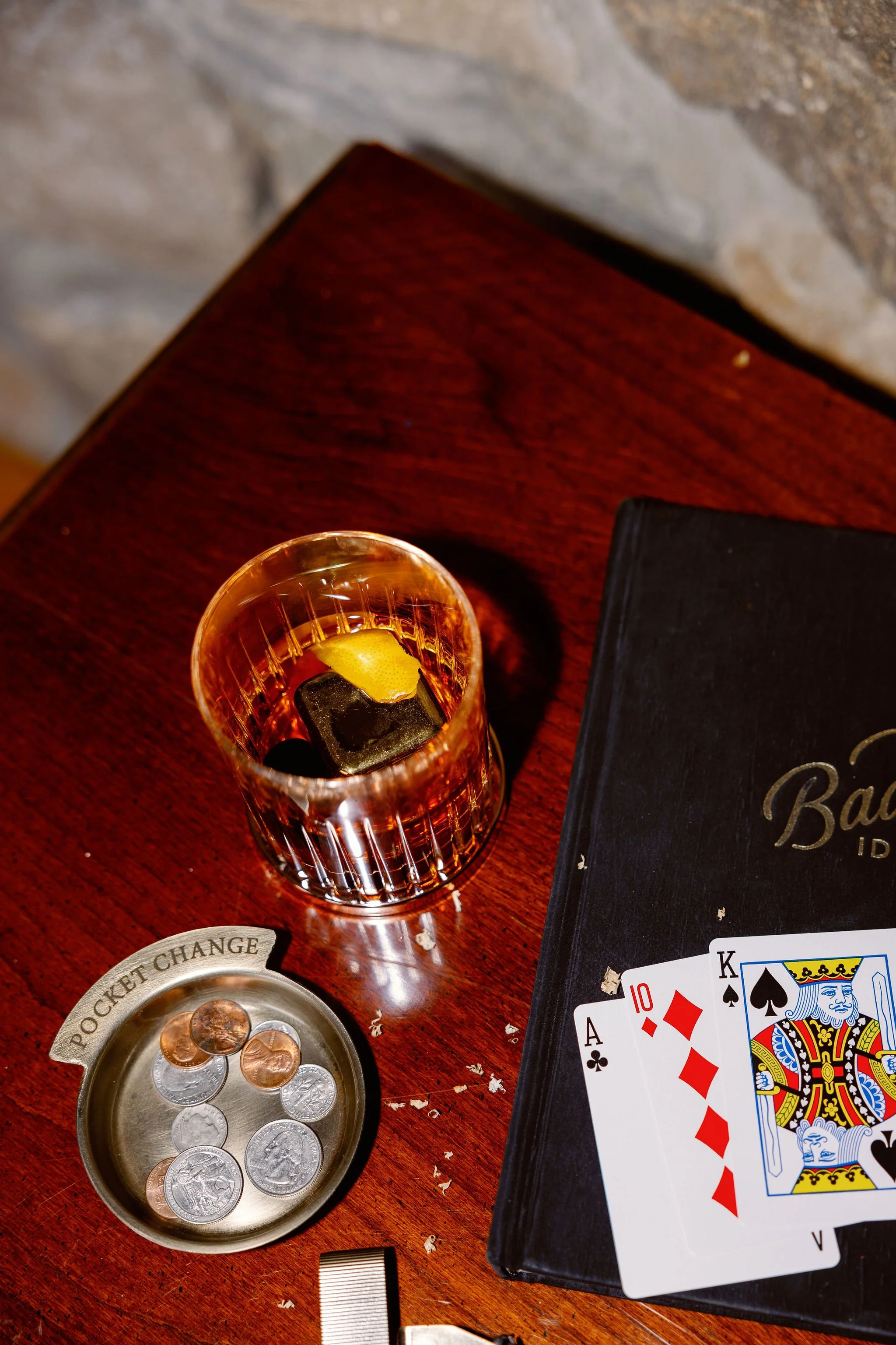 A wooden table with a glass of whiskey with ice and lemon, a black book, a coin tray labeled 'POCKET CHANGE' with coins, and a hand of playing cards showing an ace, 10, king, and queen of spades and diamonds.