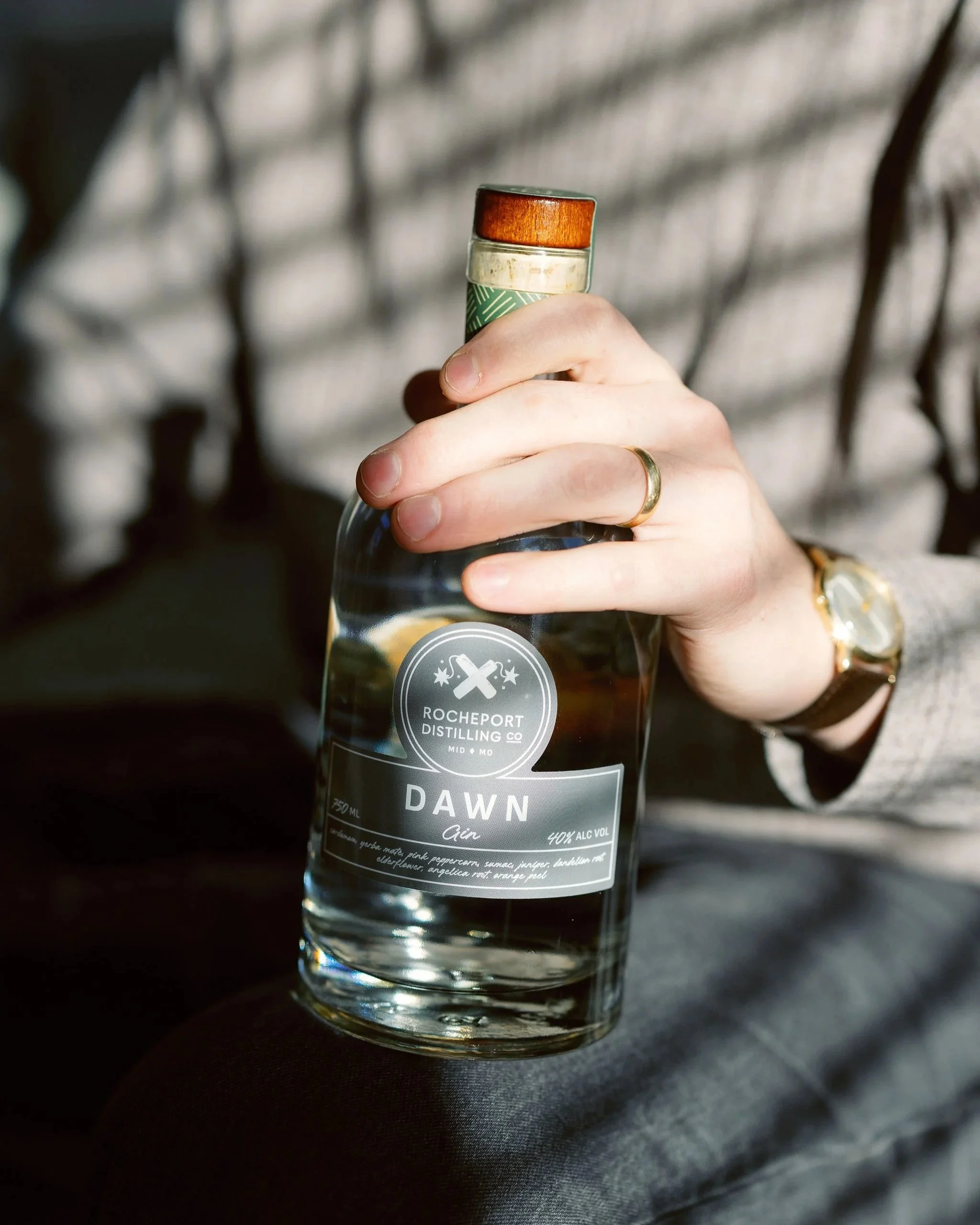 A person holding a clear bottle of alcohol with a black and white label that says 'ROCHEPORT DISTILLING CO. DAWN Air 40% ALC VOL' in front of a blurred background.