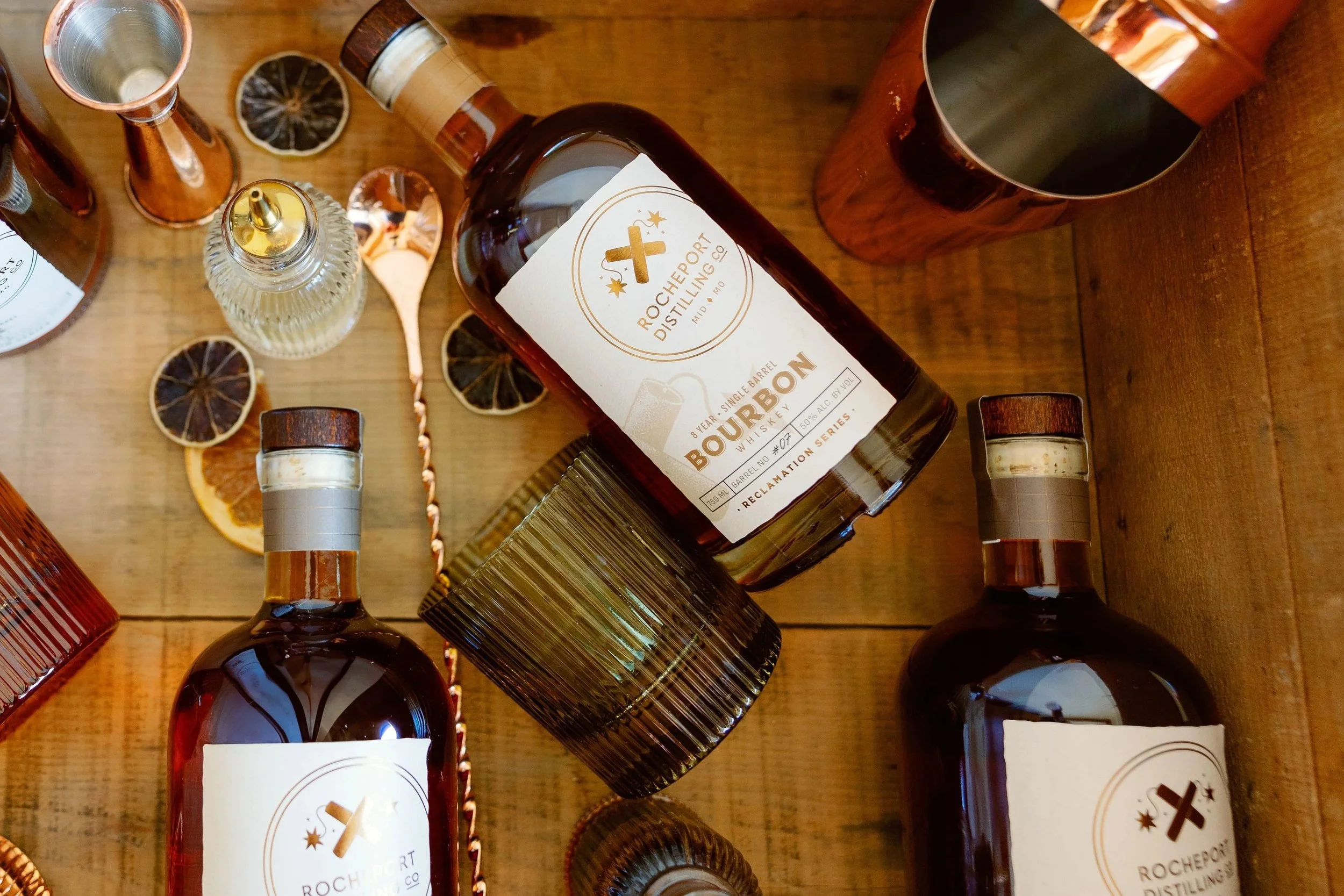 Three bottles of bourbon whiskey, Coasters, metal straws, and dried citrus slices on a wooden surface.