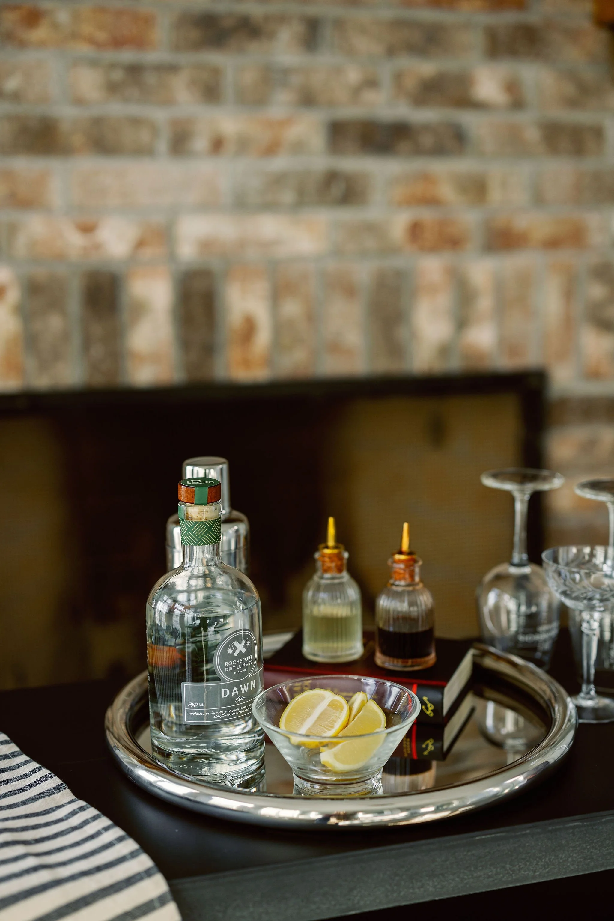 A bar tray with a bottle of vodka, lemon slices, and various bottles of bitters or mixers, set against a brick wall.