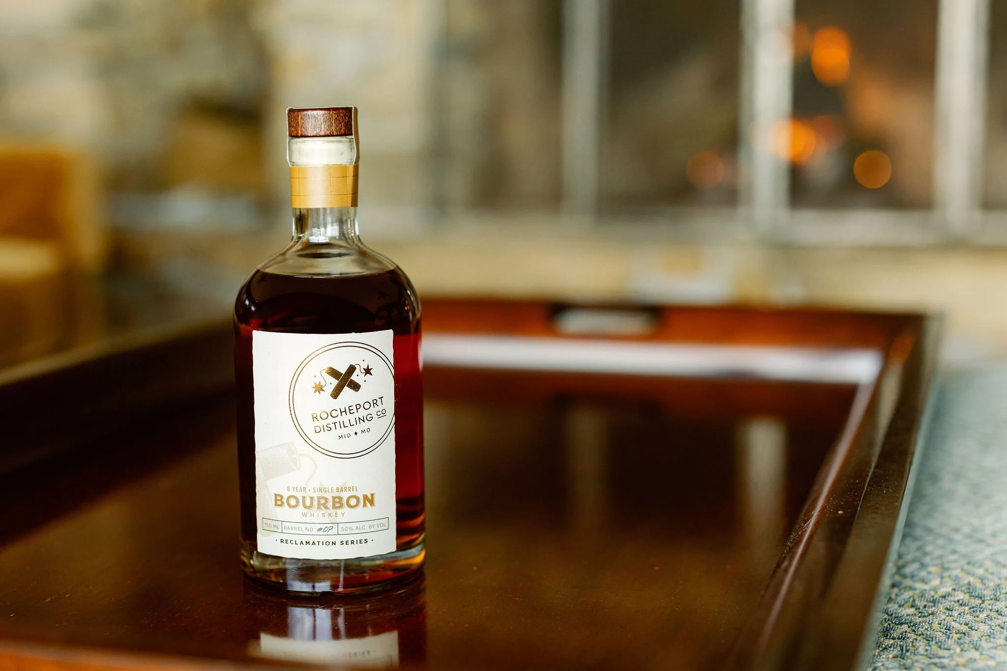 A bottle of Rocheport Distilling Co. bourbon whiskey resting on a wooden surface with a blurred background, likely in a rustic or industrial setting.