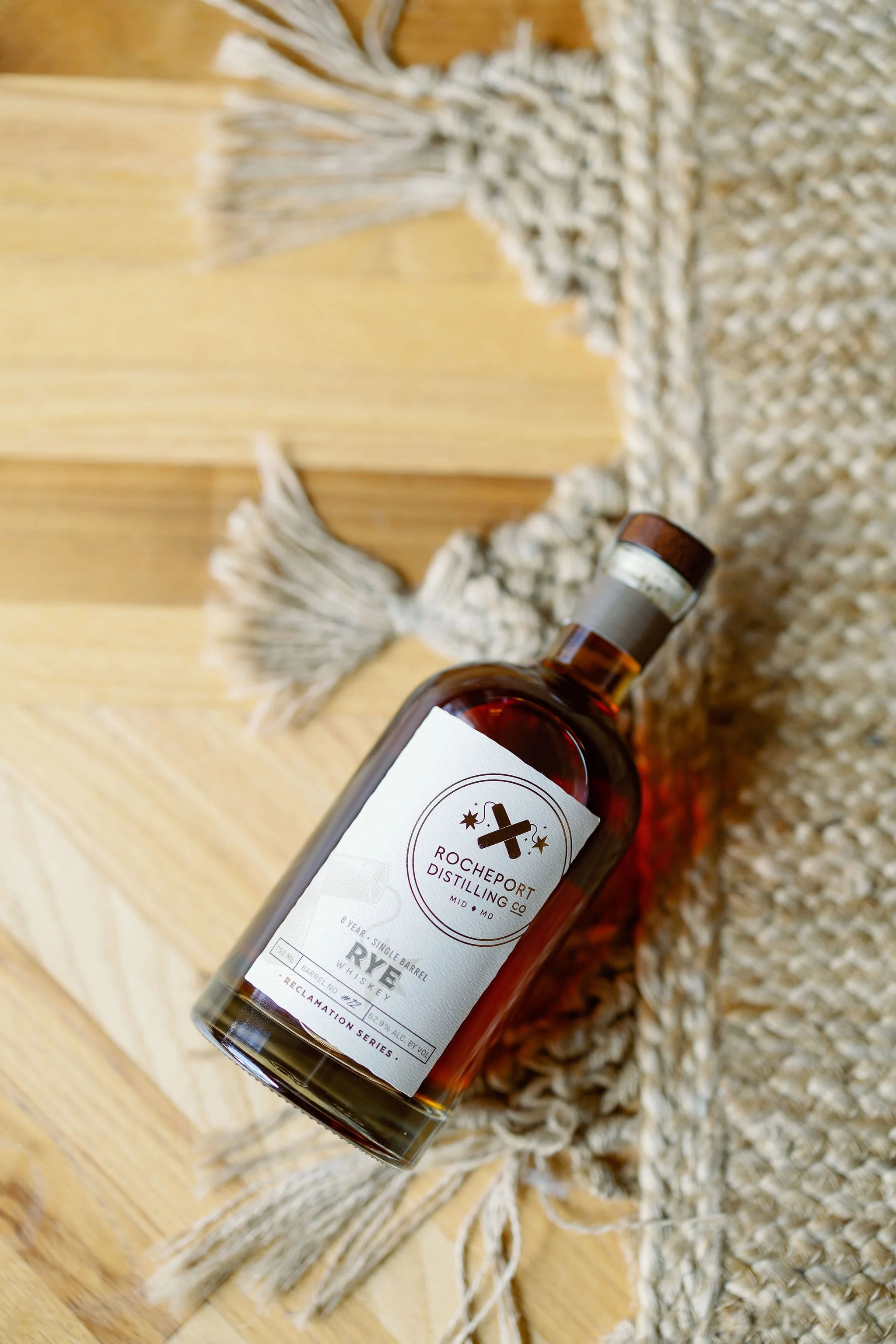 A bottle of Richeport Rye whiskey from Rocheport Distilling Co. resting on a woven blanket with tassels, placed on a wooden surface.