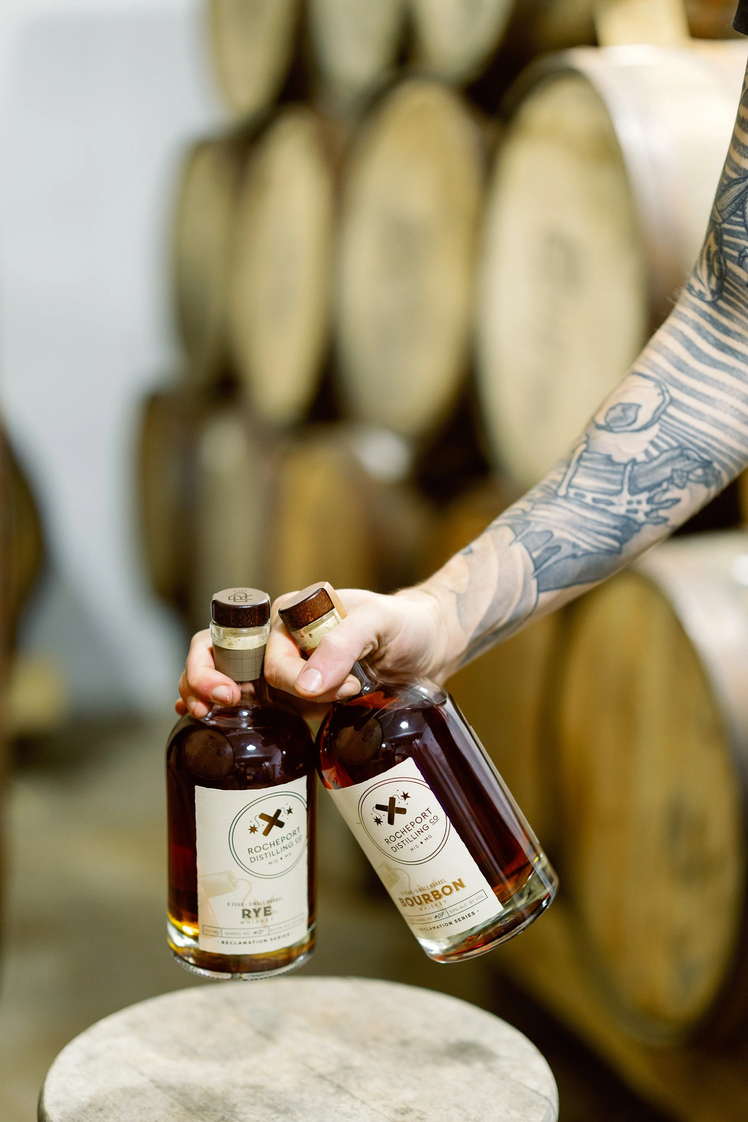 Person with tattooed arm holding two bottles of liquor in front of oak barrels.