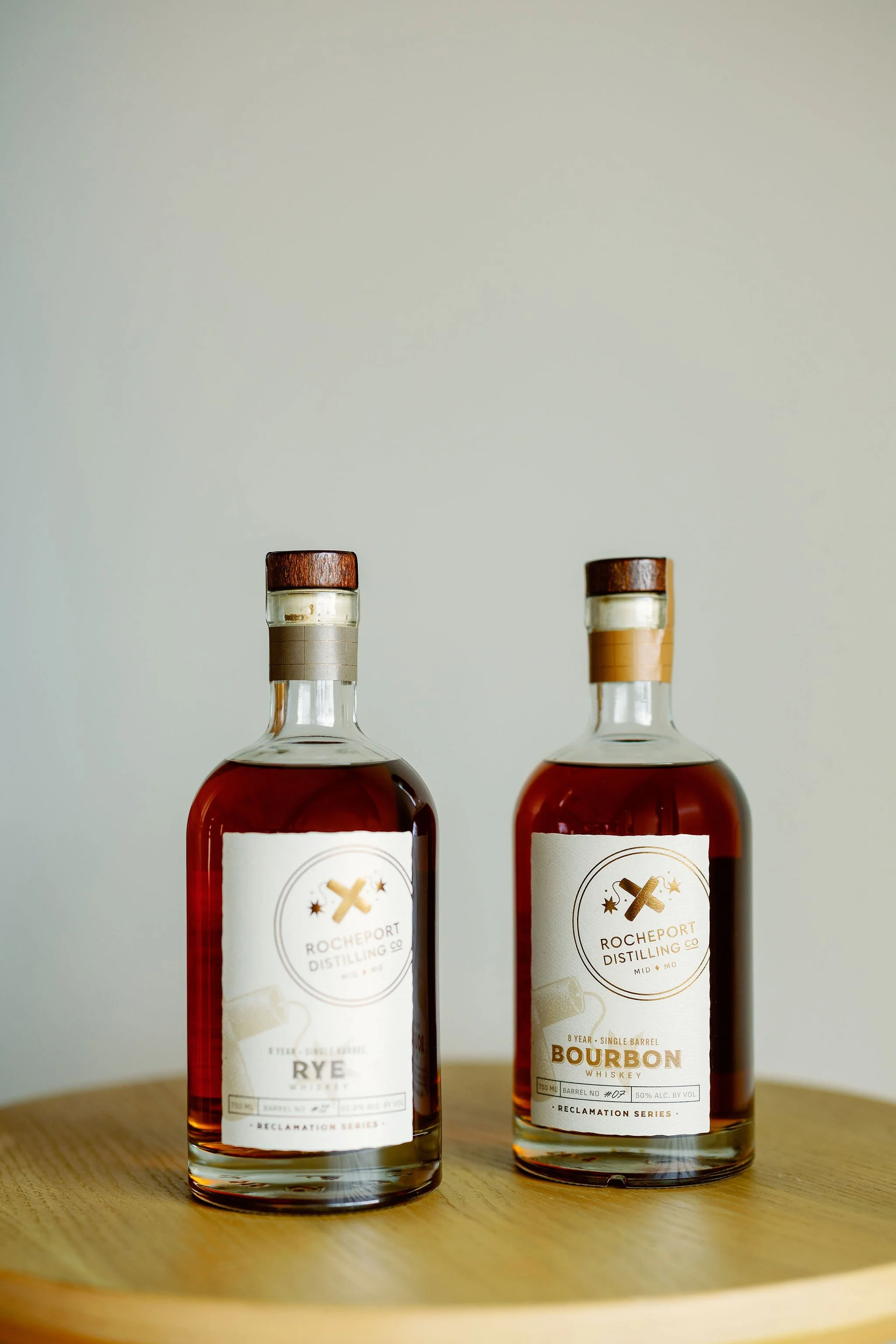 Two bottles of whiskey on a wooden surface, one labeled rye and the other bourbon, from Rocheport Distilling Co.