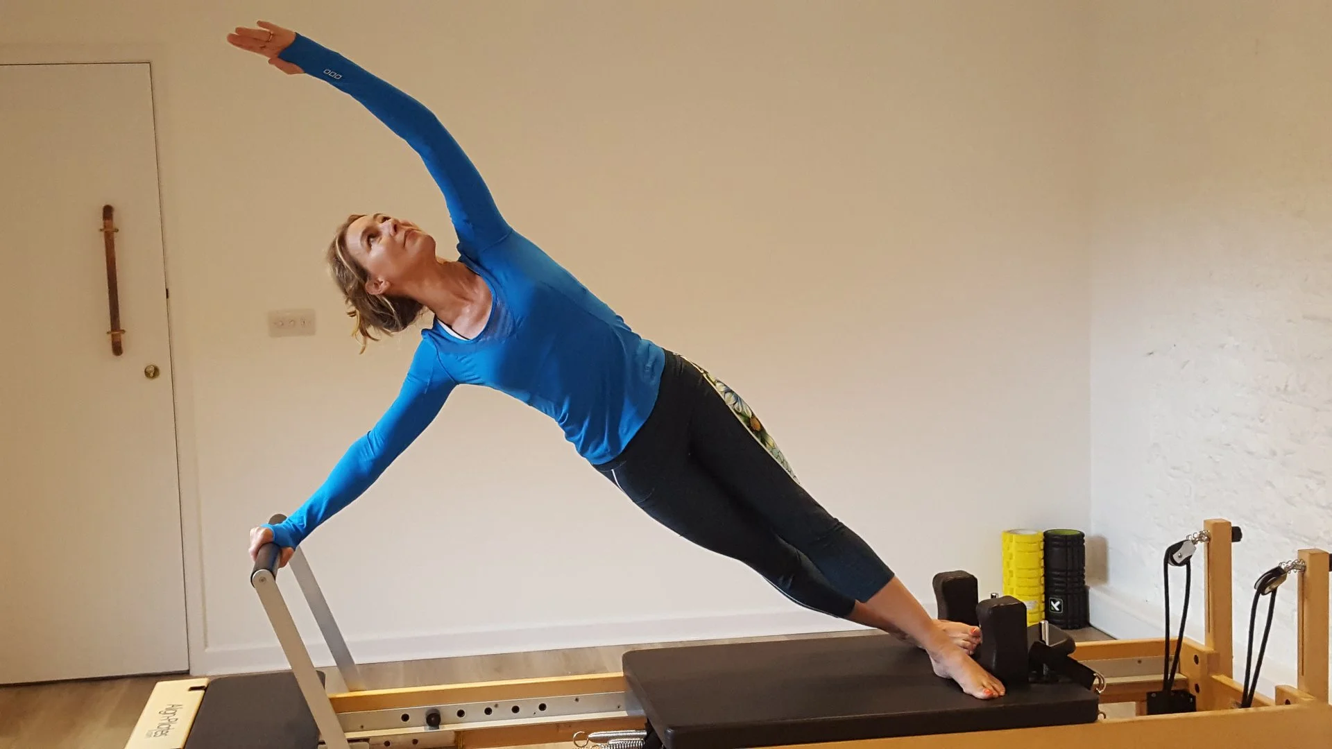 Woman practicing yoga on a Pilates reformer machine, performing a side stretch with one arm extended upward and the other holding the machine's handle, in a home gym with fitness equipment and a plain wall.