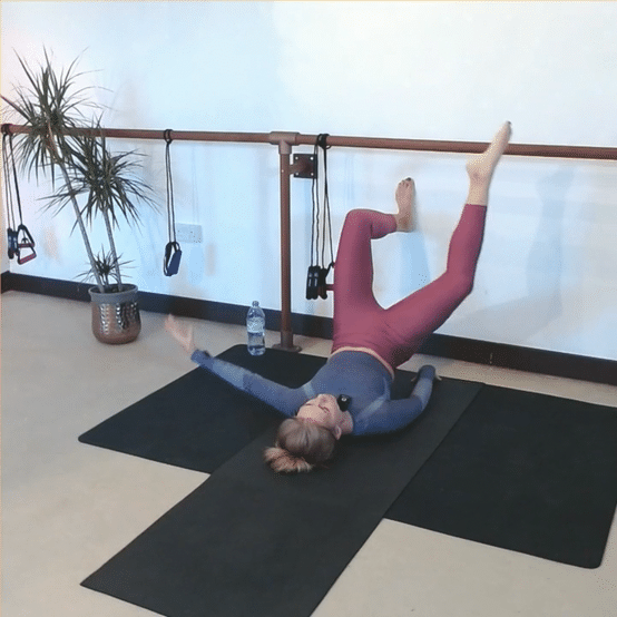 A woman lying on her back on a black exercise mat, with one leg raised and resting on a ballet barre, practicing flexibility or stretching exercises in a room with a potted plant, water bottle, and resistance bands hanging on the wall.