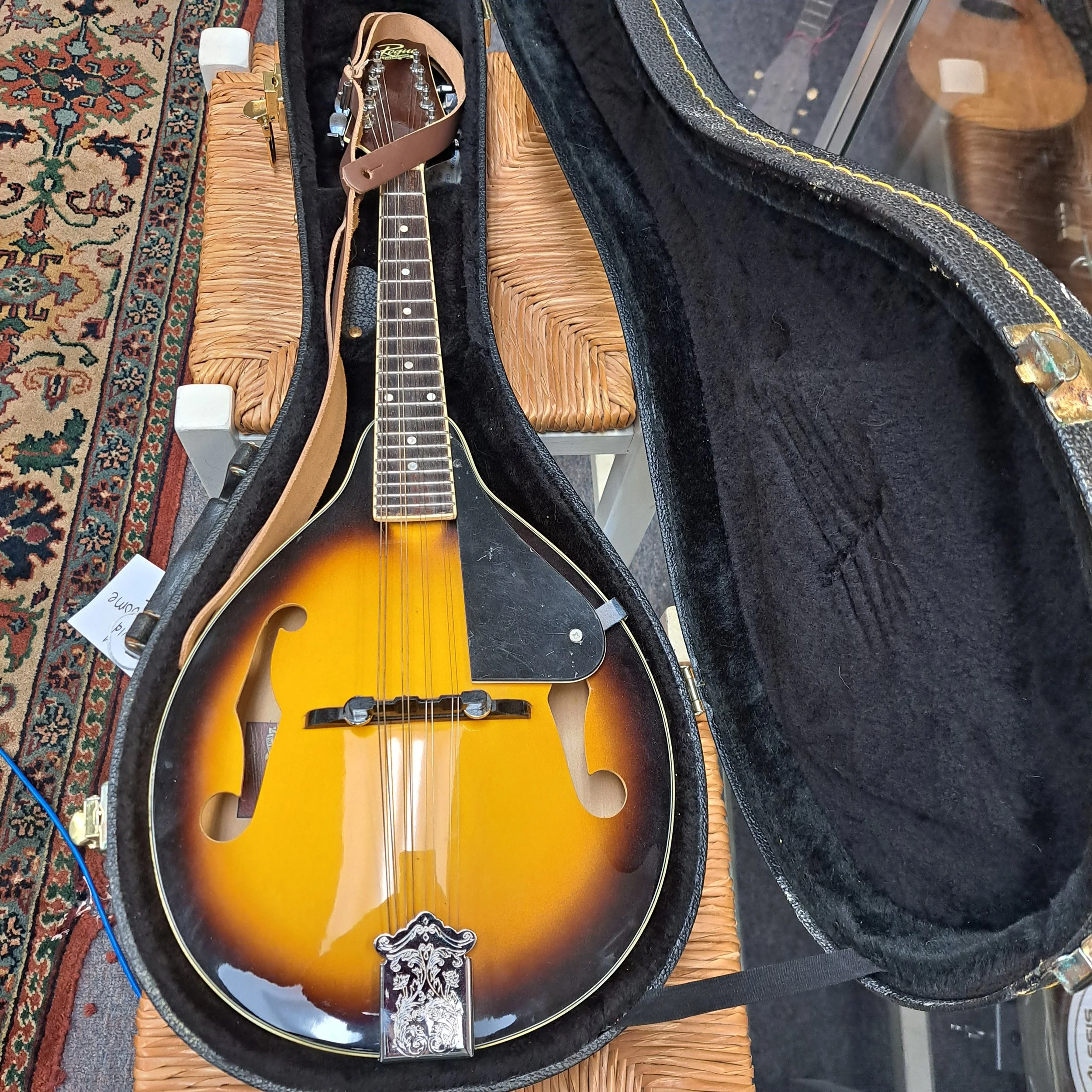 An electric guitar with a sunburst finish inside a black case lined with black velvet, resting on a woven wicker stool on a patterned rug.
