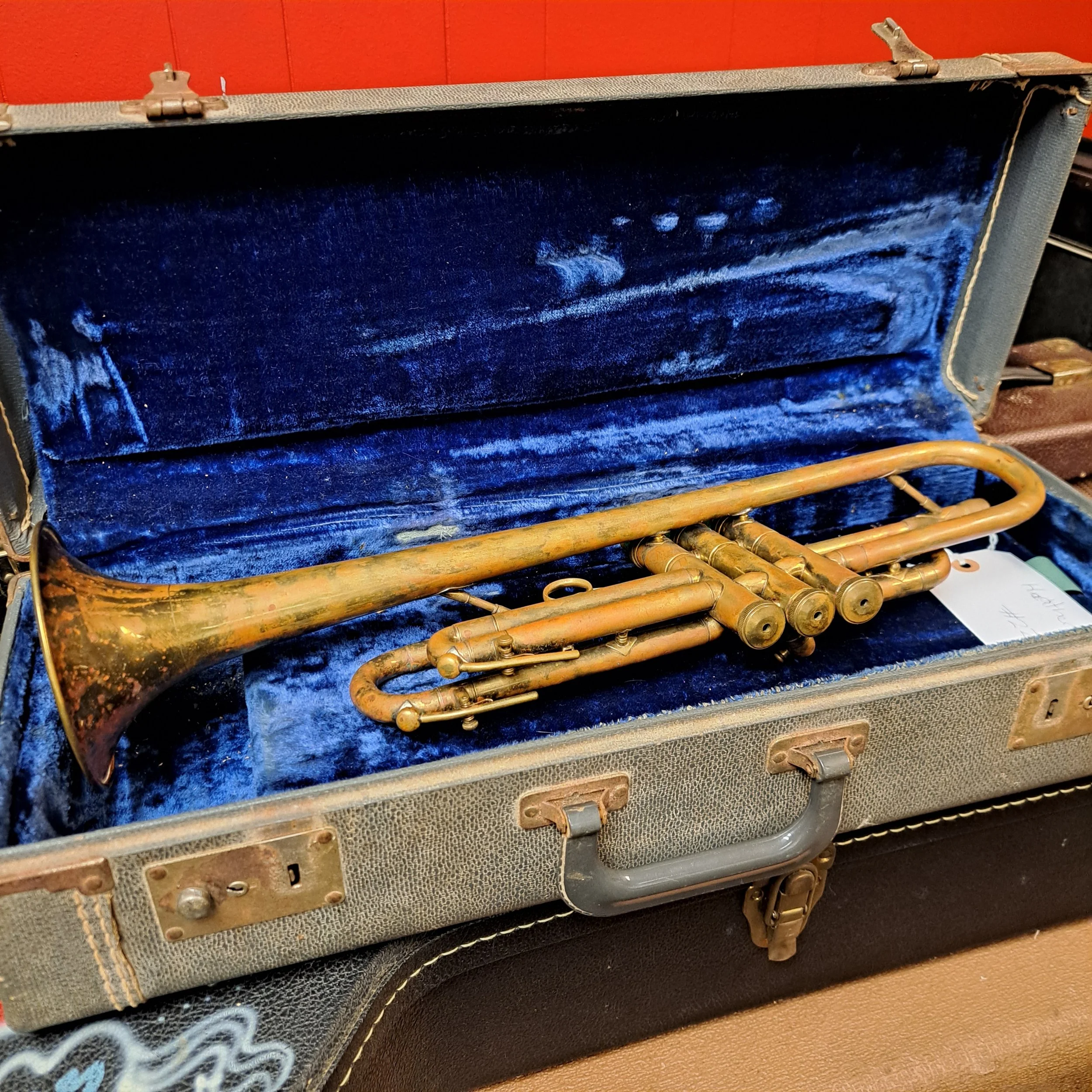 An open vintage case containing a faded, gold-colored trumpet with visible tarnish and corrosion. The case has a textured gray exterior and a plush dark blue interior lining.