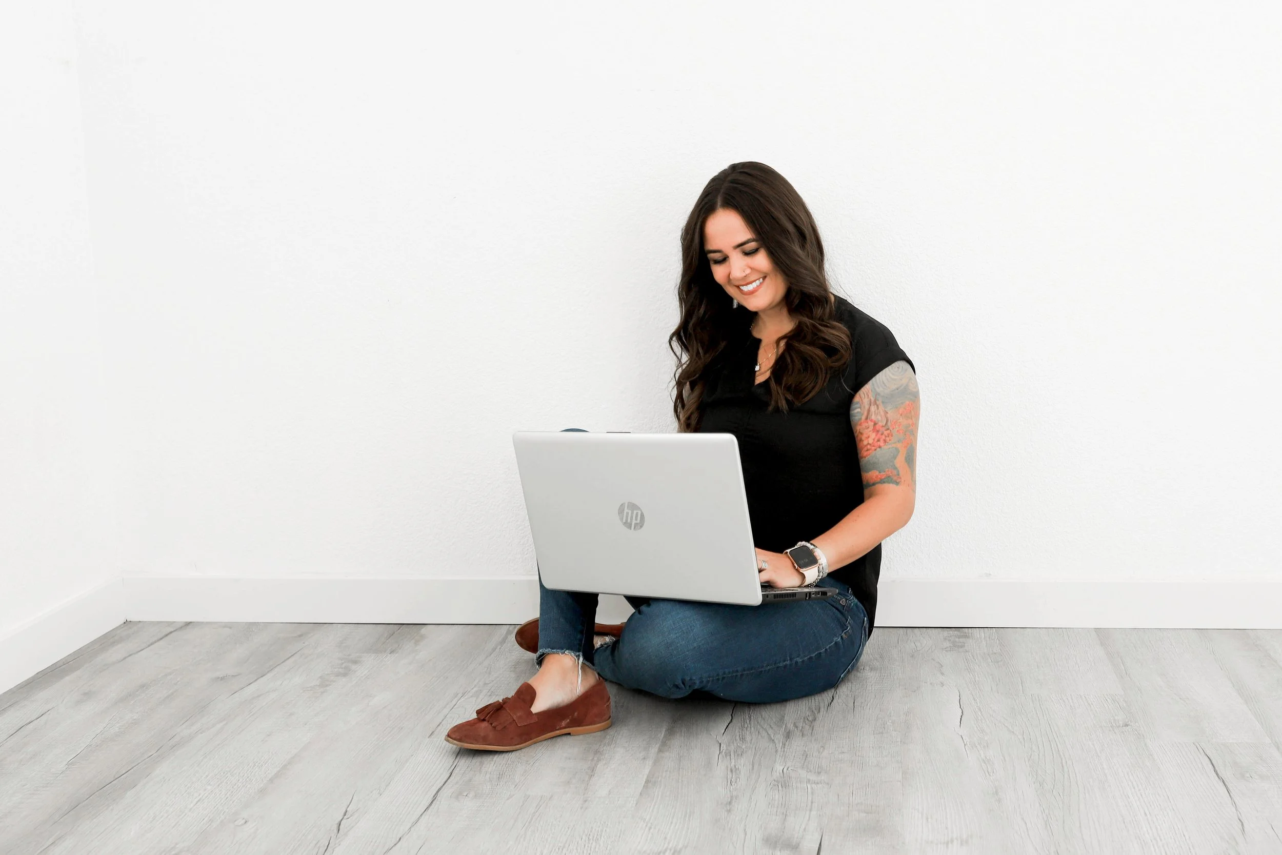 Woman sitting on floor with laptop, smiling, wearing black shirt, jeans, and brown shoes with tattoo on arm in minimalist room.
