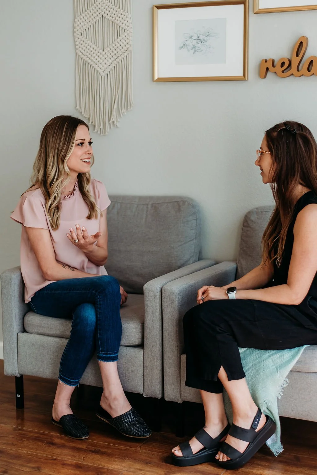 Two women sitting on gray chairs in a casual setting, engaged in a conversation. One is wearing a pink blouse and jeans, and the other is in a black outfit with an Apple Watch. The backdrop includes a macramé wall hanging and framed artwork.