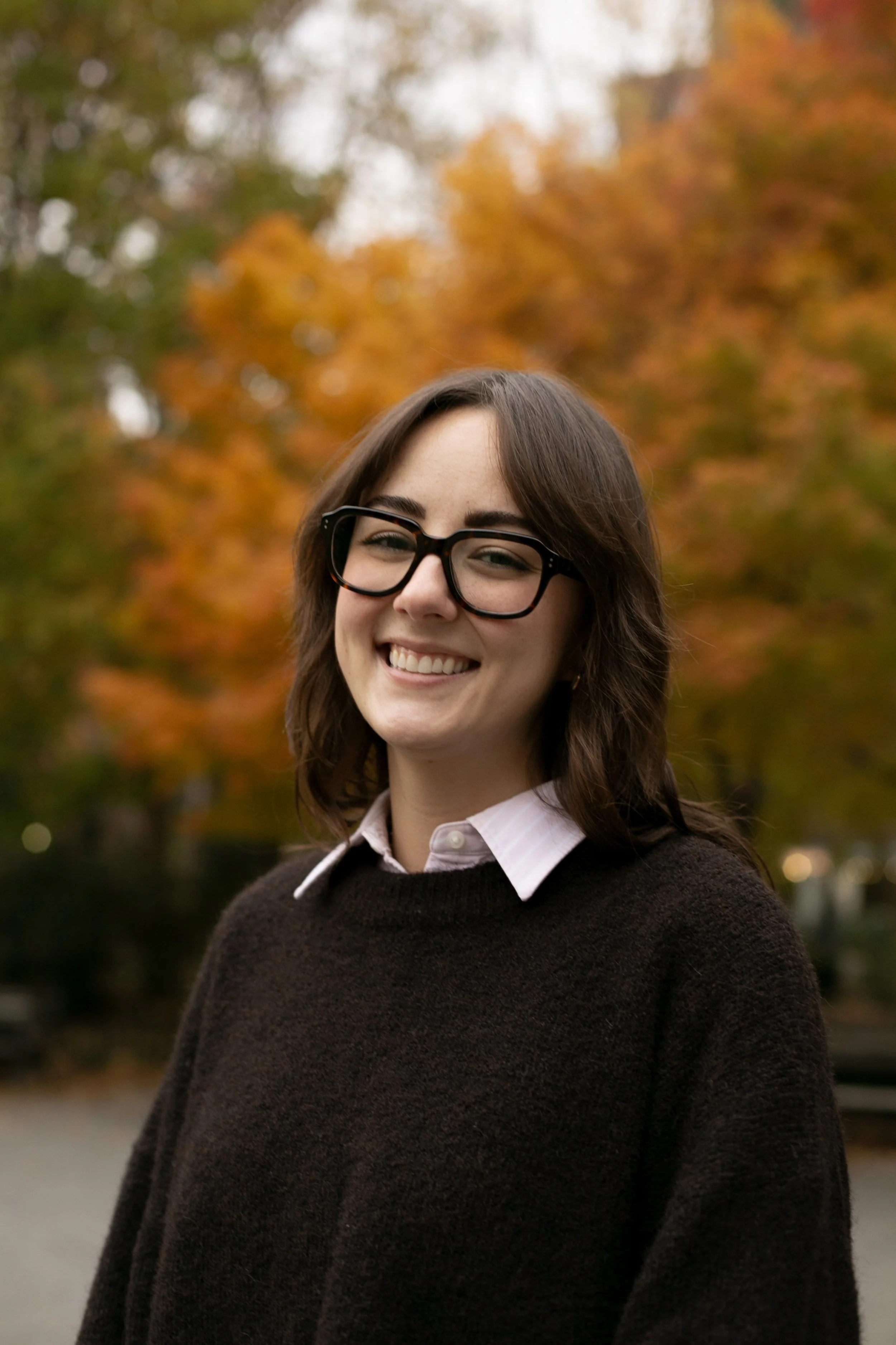 A woman with shoulder-length brown hair and black glasses smiles at the camera while standing outdoors in front of autumn trees with bright orange and yellow leaves. They’re wearing a dark sweater over a collared shirt.