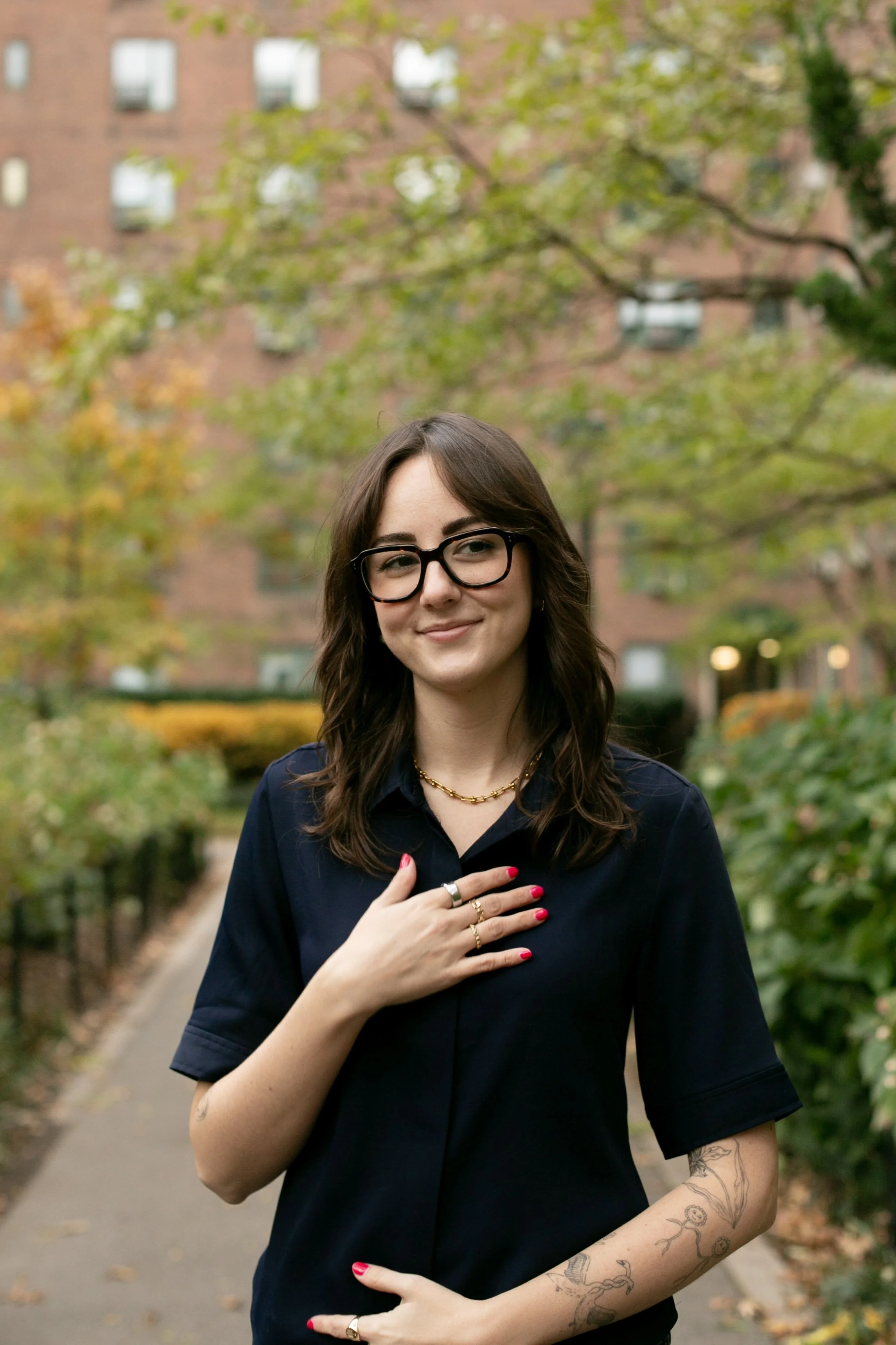 A woman with shoulder-length brown hair, glasses, and tattoos on her arm, smiling and wearing a dark blue shirt, standing in a park with green trees and a brick building in the background.
