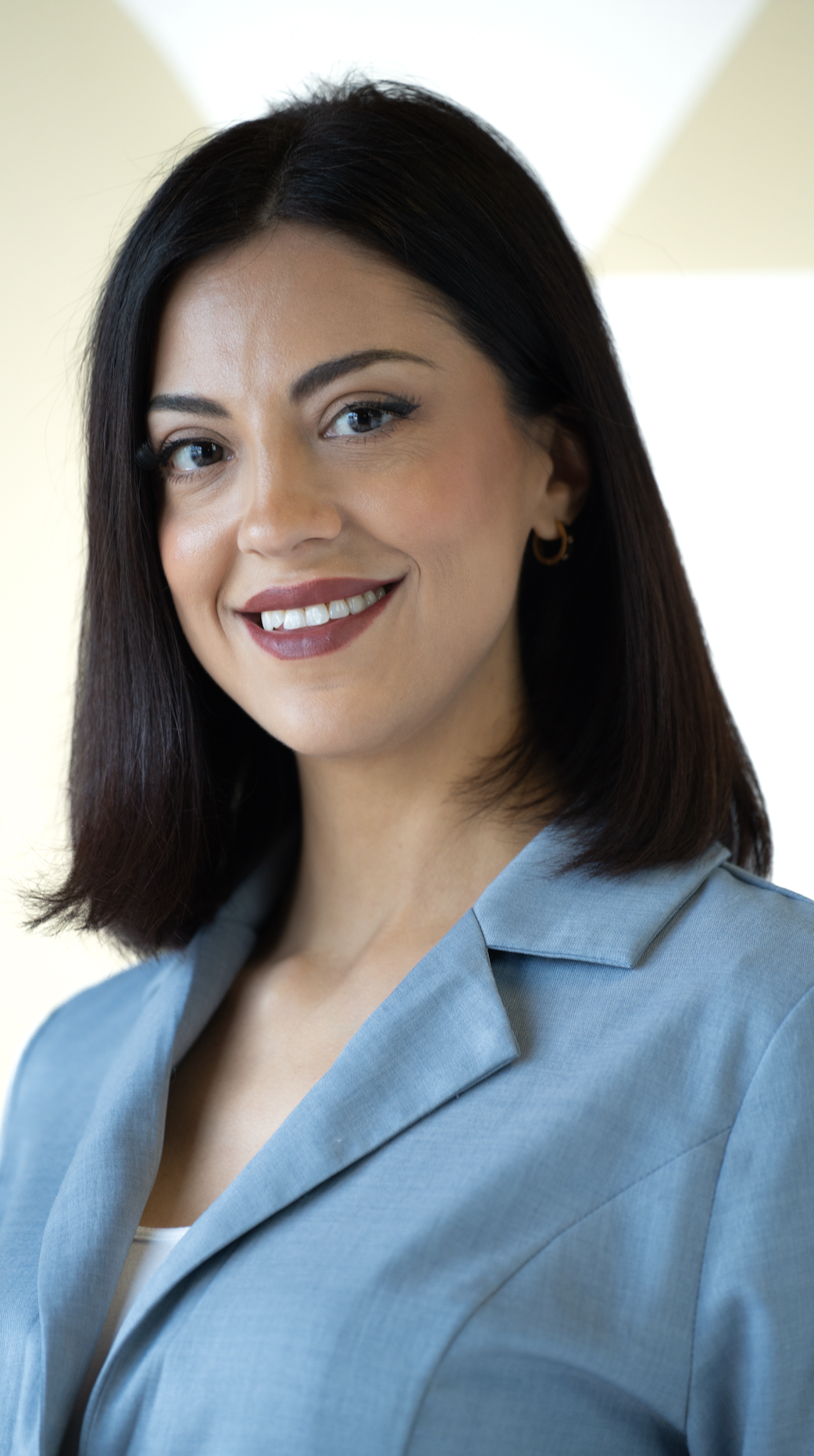 Close-up of a smiling woman with dark brown hair, wearing a light blue blazer and small gold hoop earrings.