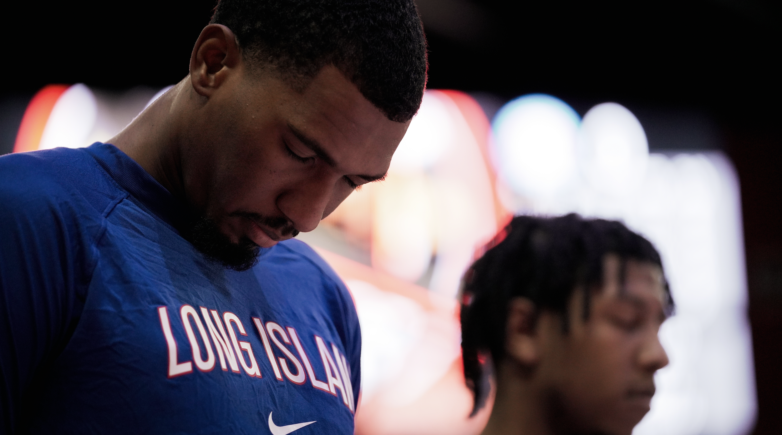 Two young men with dark skin in blue sports jerseys standing with heads bowed and eyes closed, with colorful blurred lights in the background.