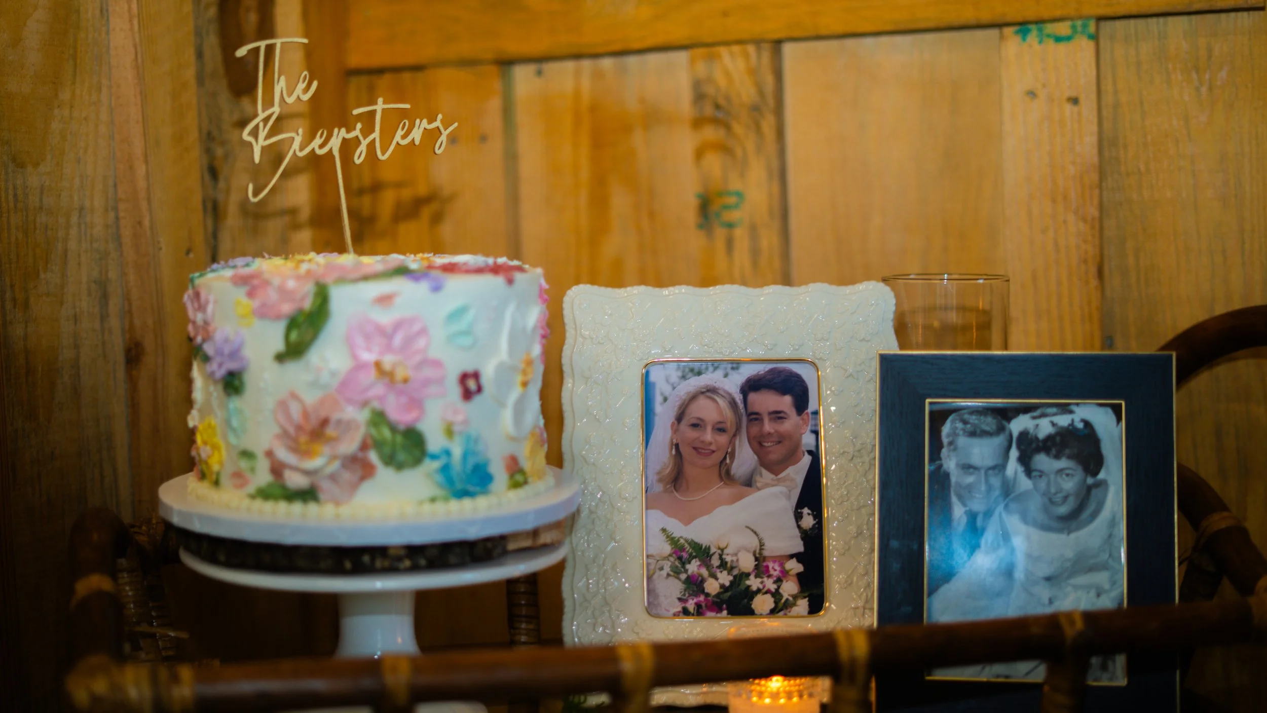 A decorated wedding cake with a topper that says 'The Bursters', and framed photographs of a couple, one in color and one in black and white, on a wooden shelf with a glass and a candle in front.