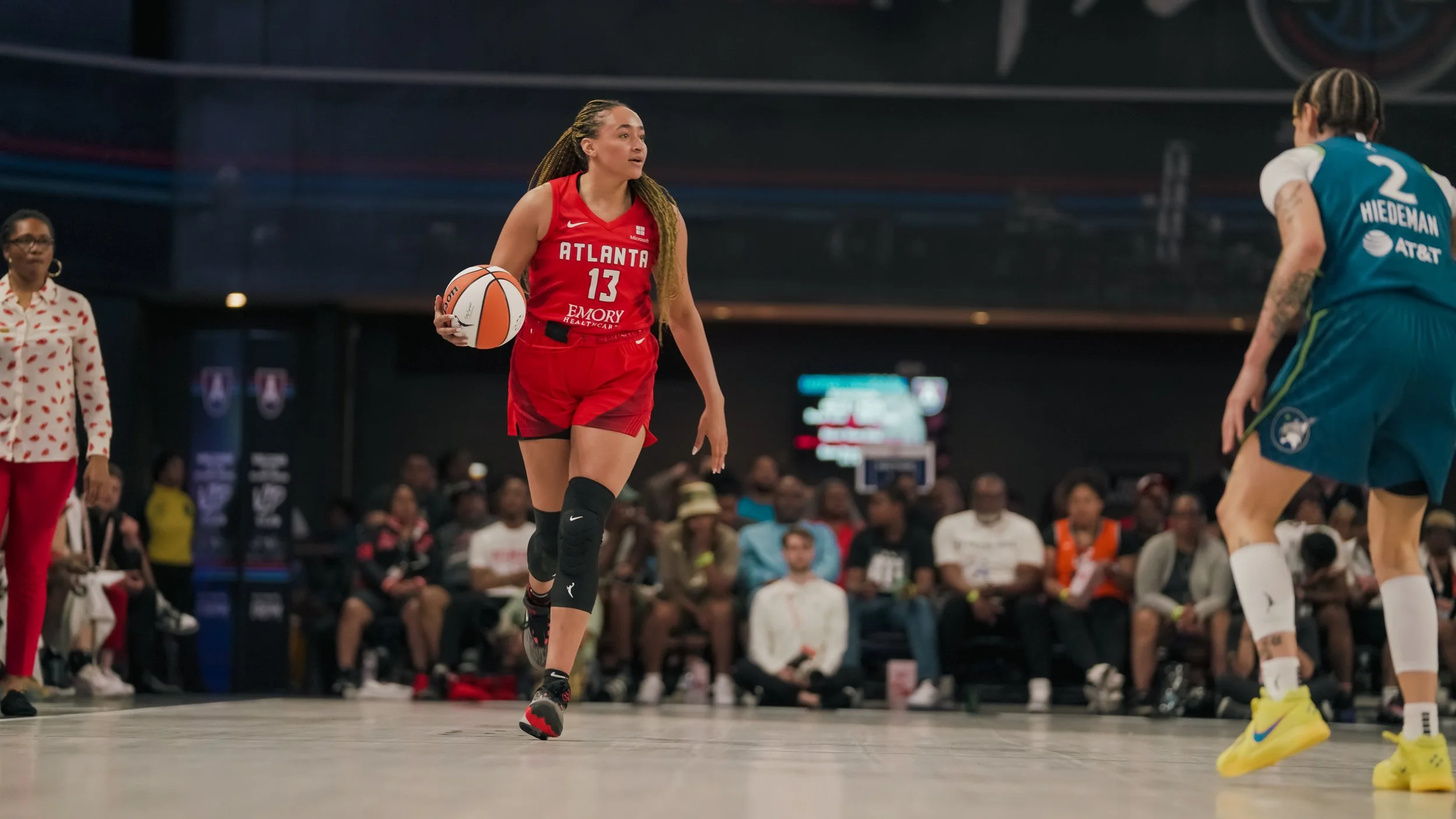 A female basketball player in a red Atlanta jersey with the number 13 holding a basketball during a game, with spectators in the background.
