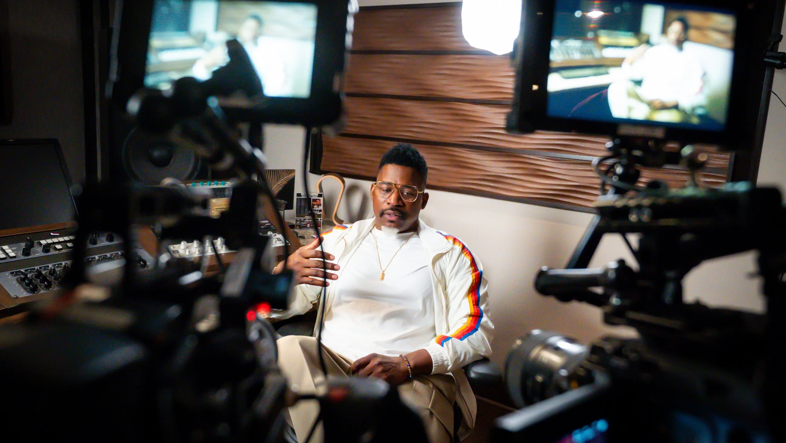 A man with glasses and a gold chain sitting in a recording studio surrounded by video cameras, with monitors showing his image and a wooden wall behind him.